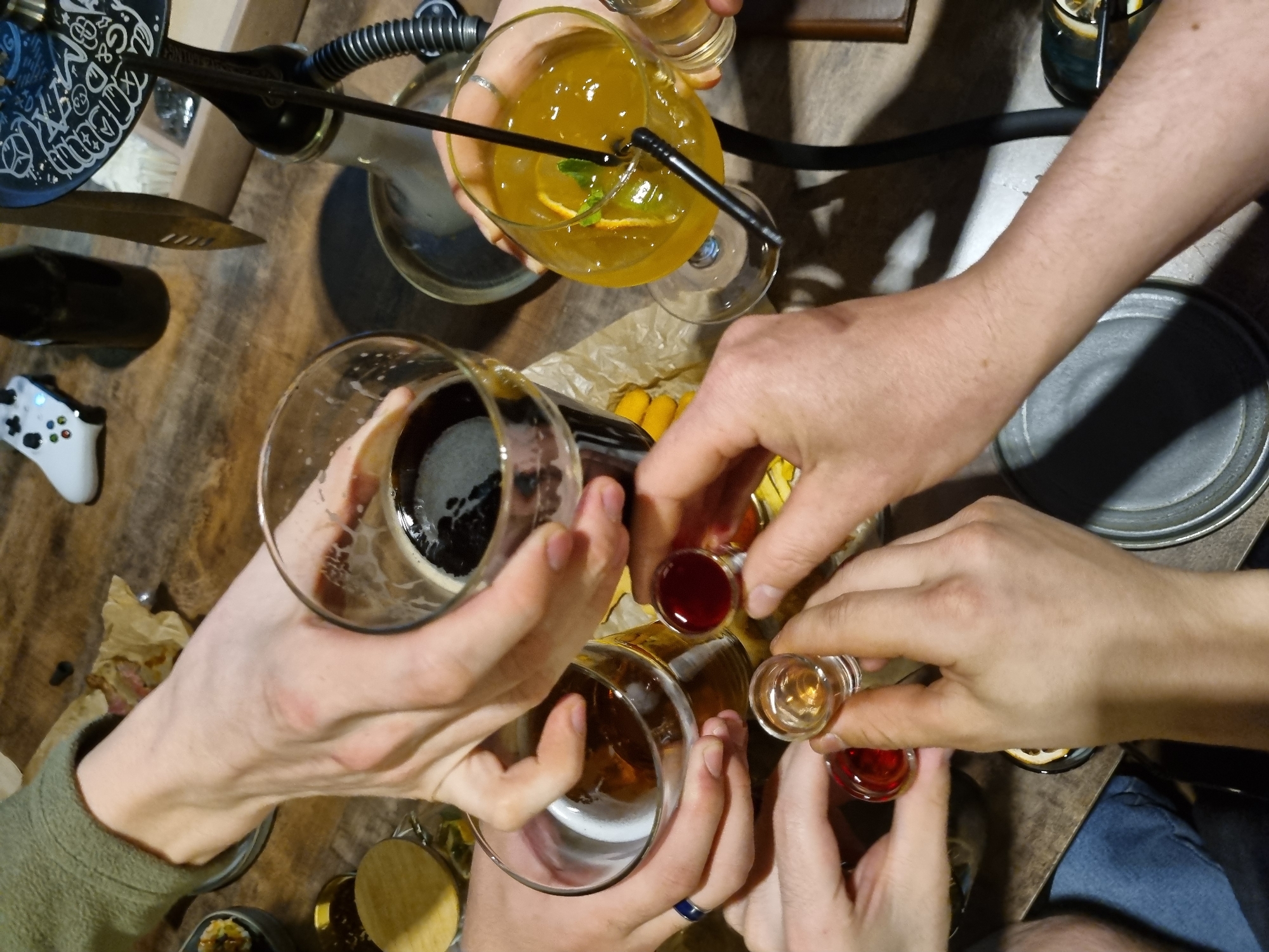 A top-down shot captures a gathering, likely a celebratory toast, on a wooden table. Hands hold glasses containing various liquids: a dark beverage in a pint glass, amber-colored beer in a stemmed glass, a yellow cocktail in a taller glass with straws, and multiple small shot glasses filled with red and clear liquids. An e-cigarette or hookah pipe rests to the upper left. A white game controller and a metal lid are also visible. The gathering seems to take place indoors, possibly in a bar or lounge setting, given the presence of alcoholic drinks and the hookah. The location is unknown.