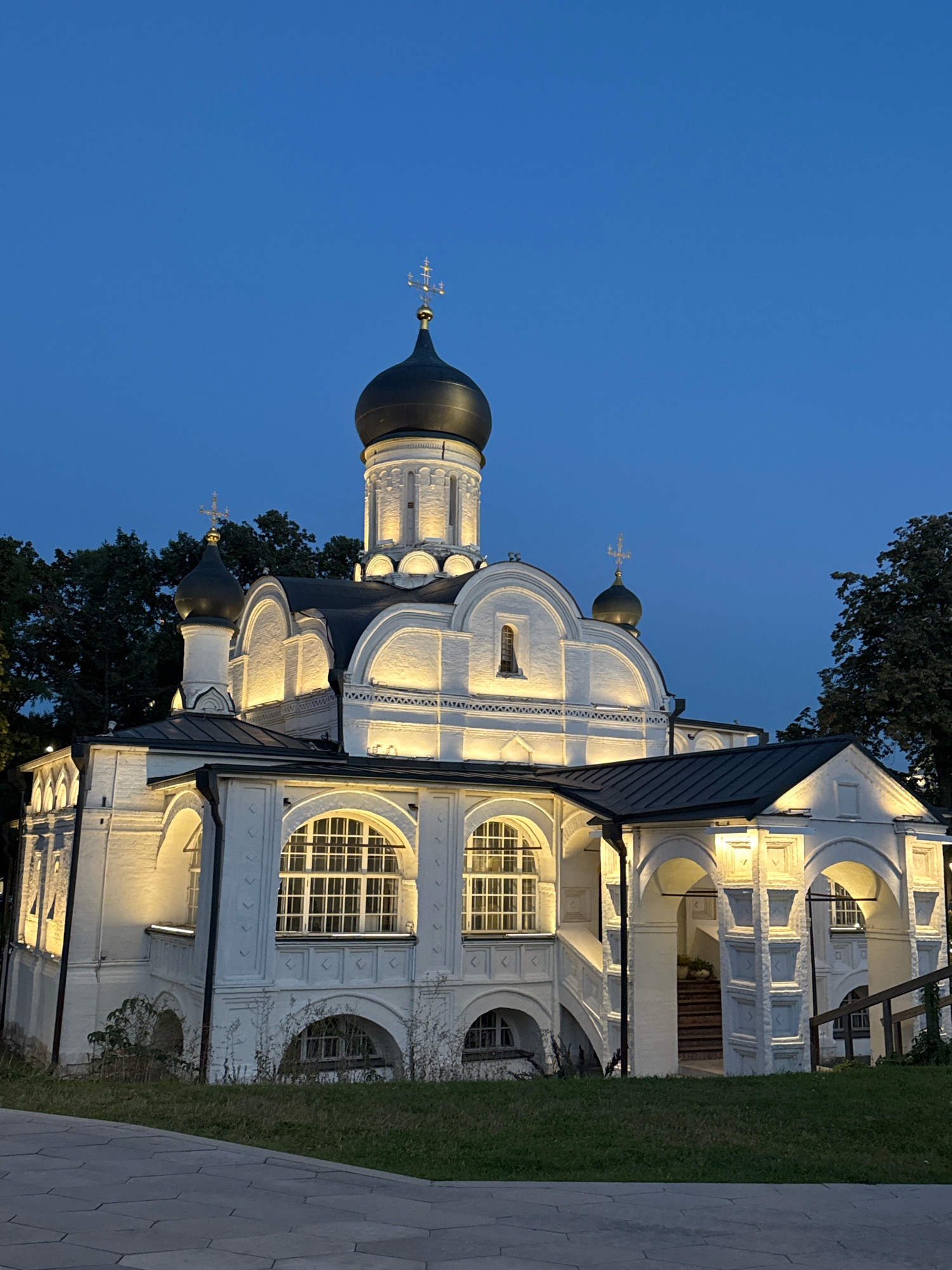 This is a nighttime, exterior, medium shot of a white, domed church in Moscow, Russia, illuminated by artificial lights. The church has multiple domes, with the largest central dome being dark and topped with a cross. Smaller dark domes are present on lower sections of the building. The architecture features arched windows with white grilles and decorative white brickwork. A dark, sloped roof covers parts of the structure. The church is surrounded by lush green grass and trees on the right side, with a paved pathway in the foreground. The sky is a deep, clear blue, characteristic of twilight or nighttime. There are no people visible in the image.