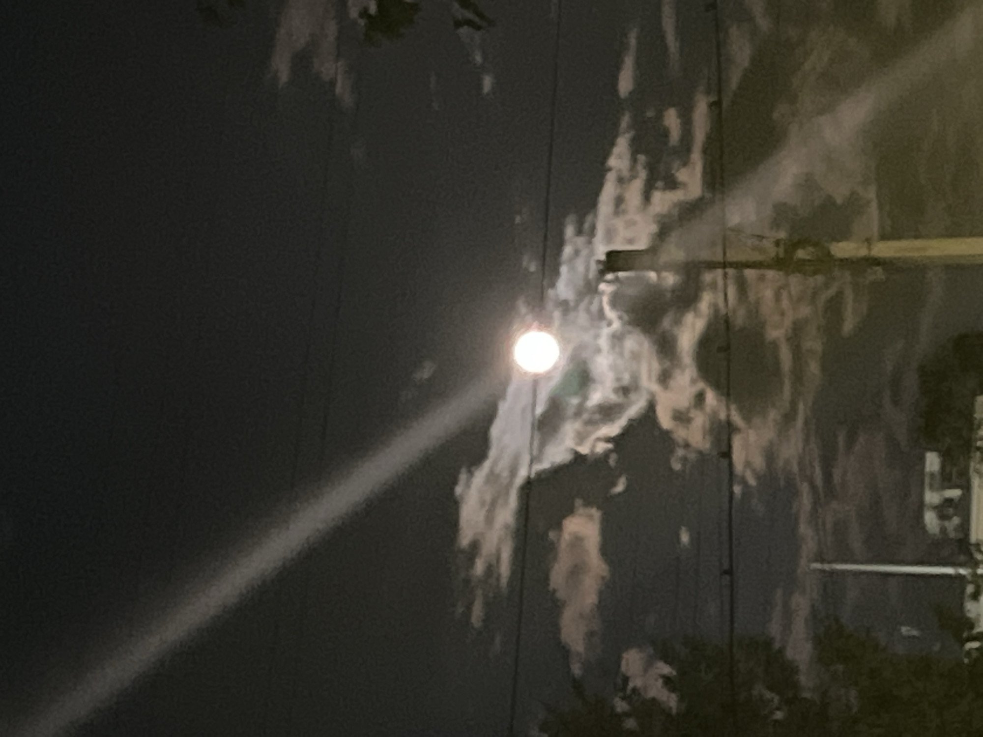 A full moon shines brightly through scattered clouds in a dark night sky. A beam of light emanates from the moon and diagonally cuts across the frame. In the foreground, a utility pole stands tall, with wires crisscrossing the sky. Below the pole, glimpses of buildings and trees can be seen. The scene appears to be set in a residential or semi-urban area.