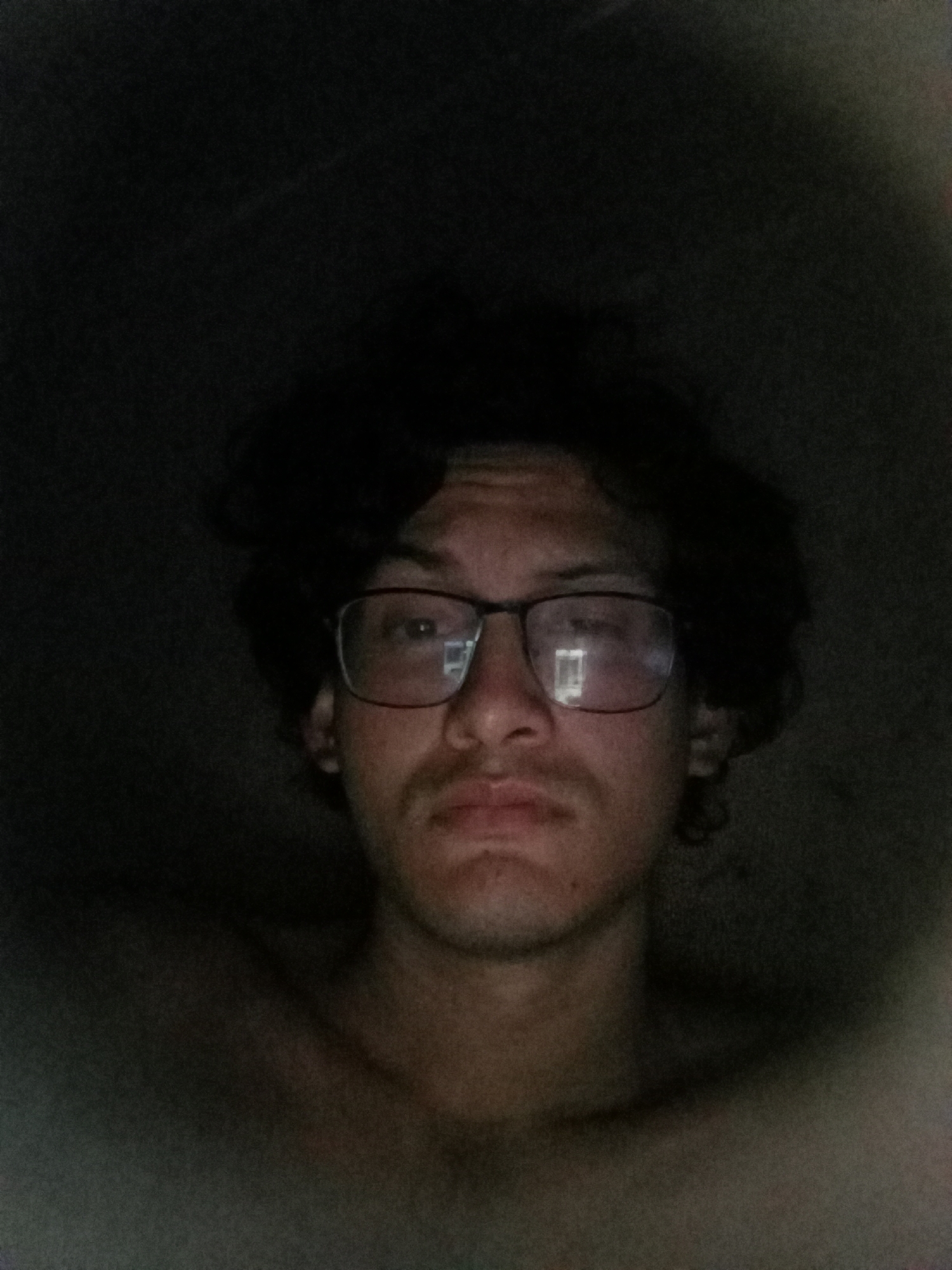 A dimly lit, close-up shot of a person wearing glasses. The person appears to be shirtless and has dark, messy hair. The background is dark and out of focus. The location in the city of Juan Griego, Venezuela cannot be ascertained from the image.