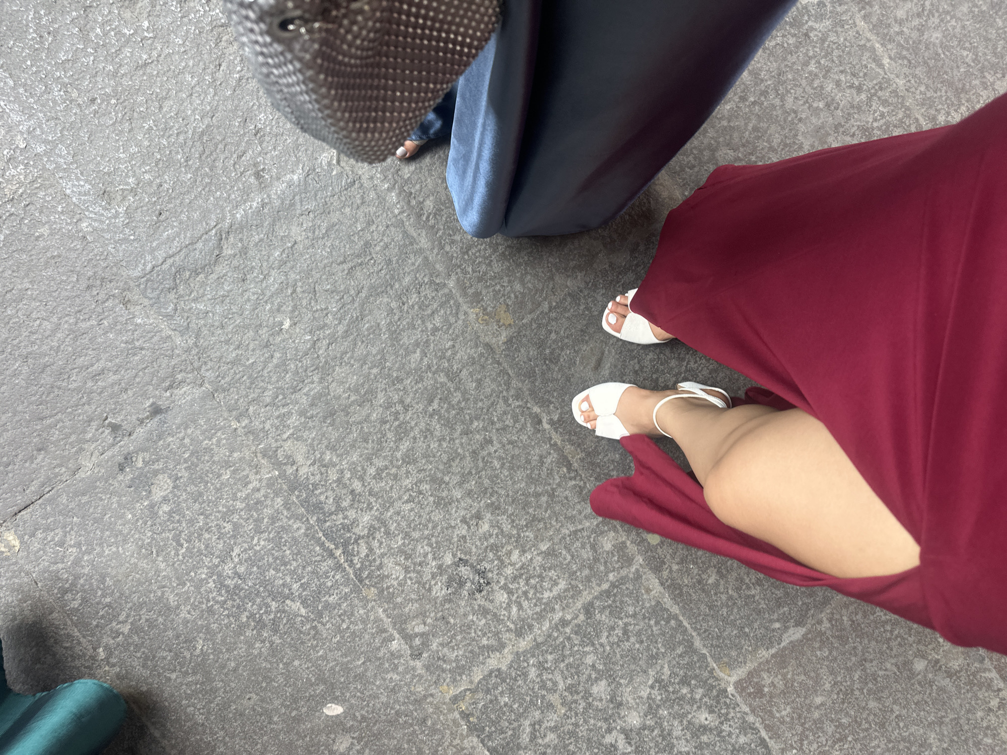 The image shows a ground-level view of a stone-paved area, with portions of three people visible. On the left, there is a person with a gray handbag, wearing dark blue attire. To the right of that person, another person is wearing a long, deep red dress and white sandals. The dress is cut in a way that exposes part of the leg. The ground is paved with light gray stones of varying sizes. The location appears to be a public space in Puebla, Mexico, due to the style of the stonework. There is a teal piece of cloth in the bottom left corner of the image.