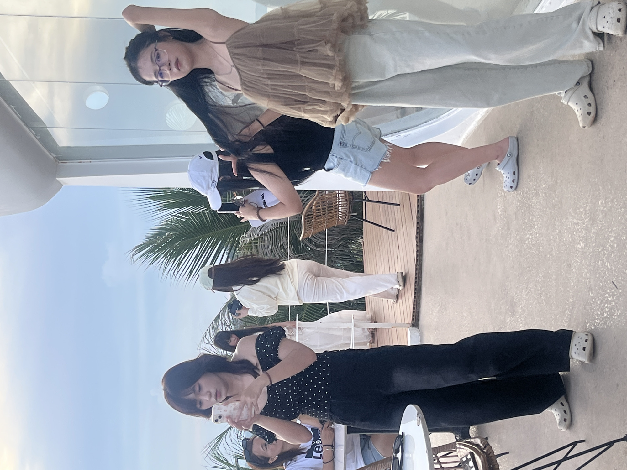 In Mũi Né, Vietnam, four young women are gathered on an outdoor elevated deck area. The woman in the foreground on the left is wearing a black off-the-shoulder top with white polka dots and black wide-leg pants. She is holding a smartphone in her left hand and looking at it. To her right, partially obscured, is another woman wearing a white dress and a light-colored hat. Further to the right, a woman in a white baseball cap and black tank top is wearing denim shorts. She is holding a camera. The woman on the far right is wearing a tan, tiered sleeveless top and light-colored pants. She has her hand raised to her hair. The environment appears to be a restaurant or cafe with wooden decking and a railing, overlooking a tropical landscape with palm trees. The sky is partly cloudy.