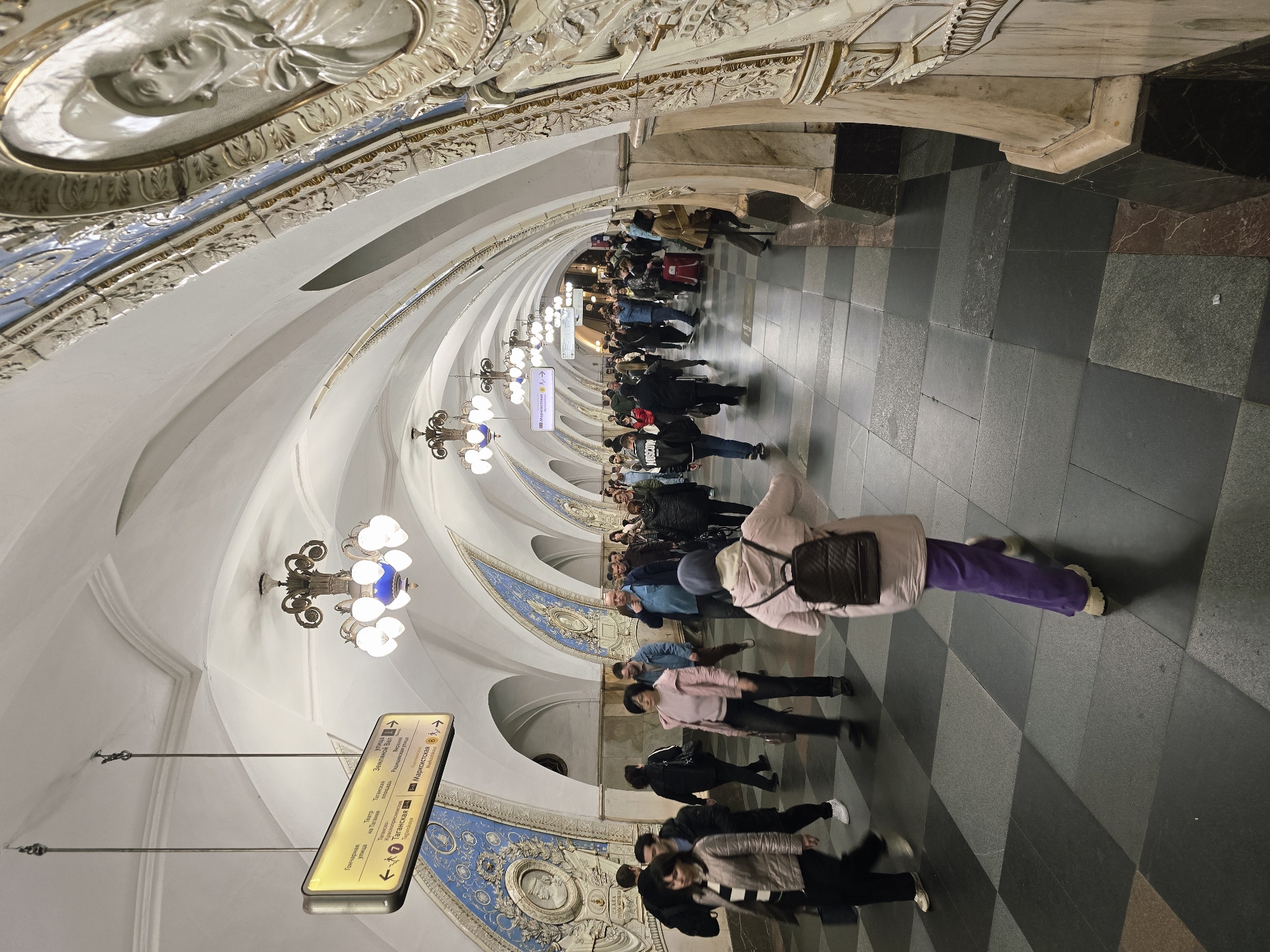 Here is a detailed description of the image:
**Setting:** The image is taken in what appears to be a subway station. The architecture is notable for its ornate, vaulted ceilings adorned with decorative details, including bas-reliefs, moldings, and chandeliers. The floor is tiled in a checkered pattern with gray and black tones.
**People:** There are numerous people visible in the image, suggesting a crowded public space. Most are blurred, indicating movement. They are dressed in casual attire. A prominent figure is a person in the foreground wearing a light-colored jacket and purple pants, walking toward the camera. Many of the people are walking in the same direction, indicating a flow of commuters.
**Objects:**
- Hanging illuminated signs with directional information are visible.
- Chandeliers hang from the arched ceiling.
- Backpack being worn by the person in the foreground.
**Actions:** The primary action is people walking through the subway station.
**Environment:** The subway station has an air of grandness due to its architectural style. The lighting is generally bright, with some areas appearing darker due to the perspective and angle of the photo.