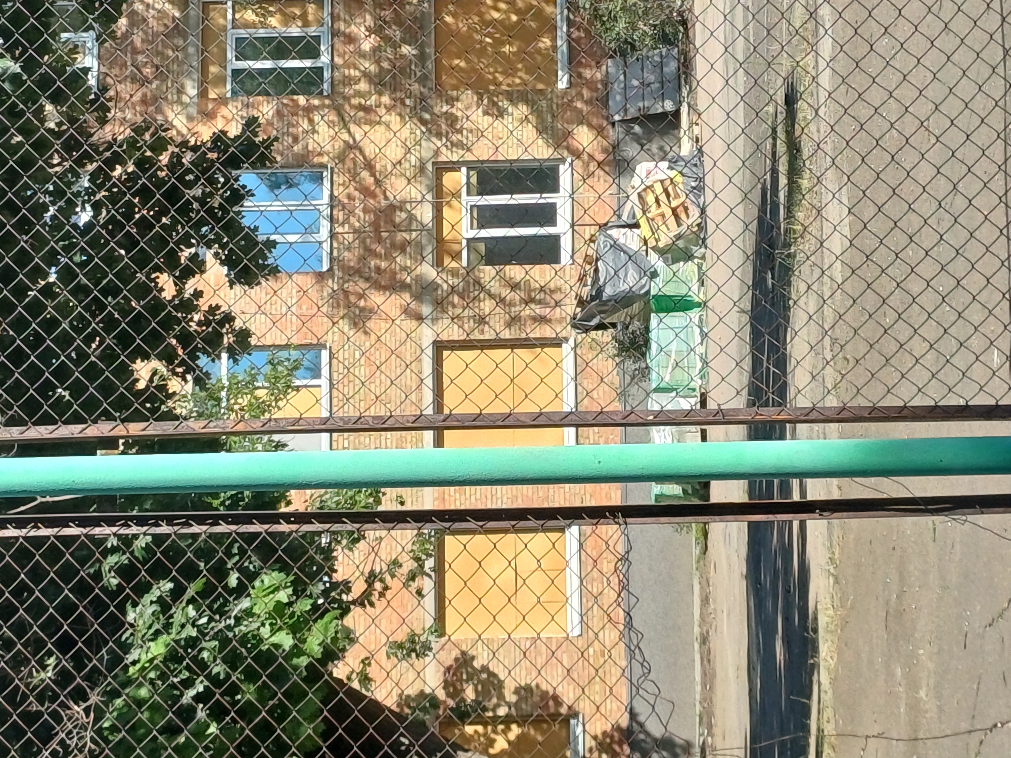 This is a photograph taken through a chain-link fence of a brick building in Kyiv, Ukraine. The building has multiple windows, some of which are covered with yellow material. There is a green pole in the foreground, part of the fence. Piles of construction materials, including plastic sheeting and wooden pallets, are visible near the base of the building. Trees with green foliage are casting shadows on the building. The ground in front of the building appears to be paved.
