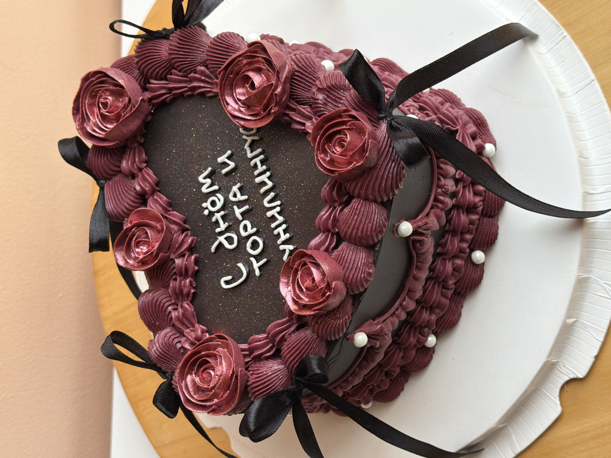 A heart-shaped cake with dark brown frosting is adorned with maroon piped roses and small white sugar pearls. Black satin ribbons are tied around the edges of the cake. In white icing, vertical text is written on the cake. The cake sits on a white corrugated cake board which is itself on a light brown surface. The location or city is not visually discernable in this image.