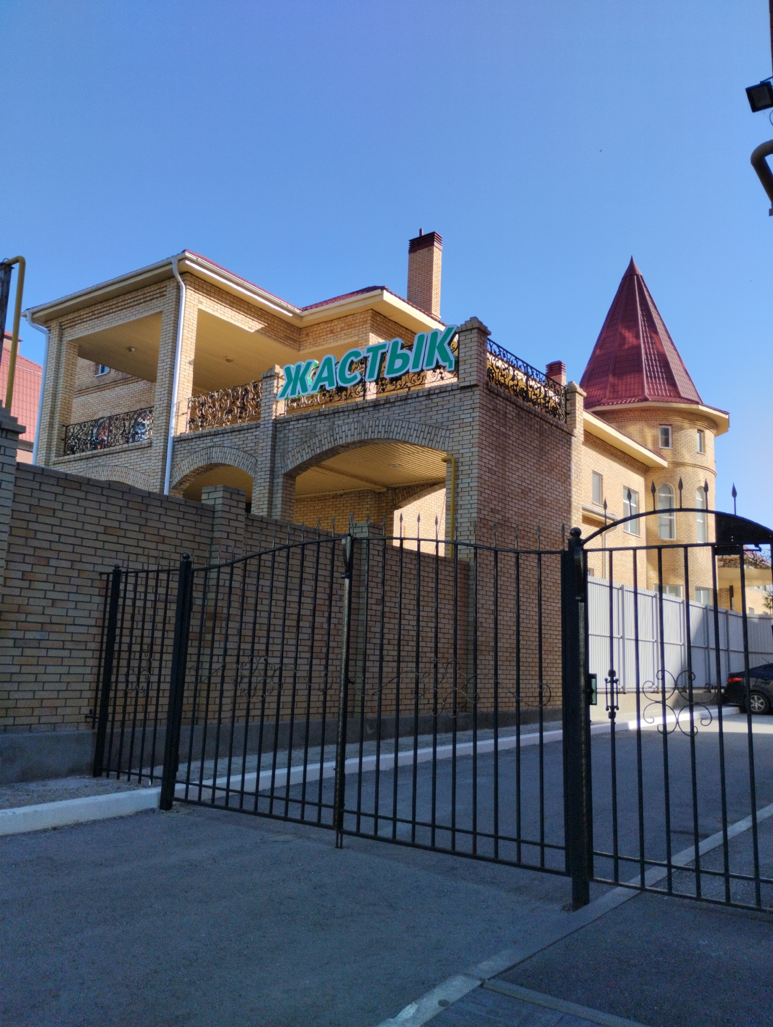The image shows a two-story building with a red tiled roof and light brown brick exterior. A sign with green letters, partially obscured by the roofline, reads "ЖАСТЫҚ". A black metal fence is in the foreground, with a paved driveway behind it. The sky is clear and blue. The building appears to be a business or residential complex. The sign likely indicates the name of the establishment or complex. The overall scene suggests a sunny day in an urban or suburban environment. This building is located in Kostanay, Kazakhstan.