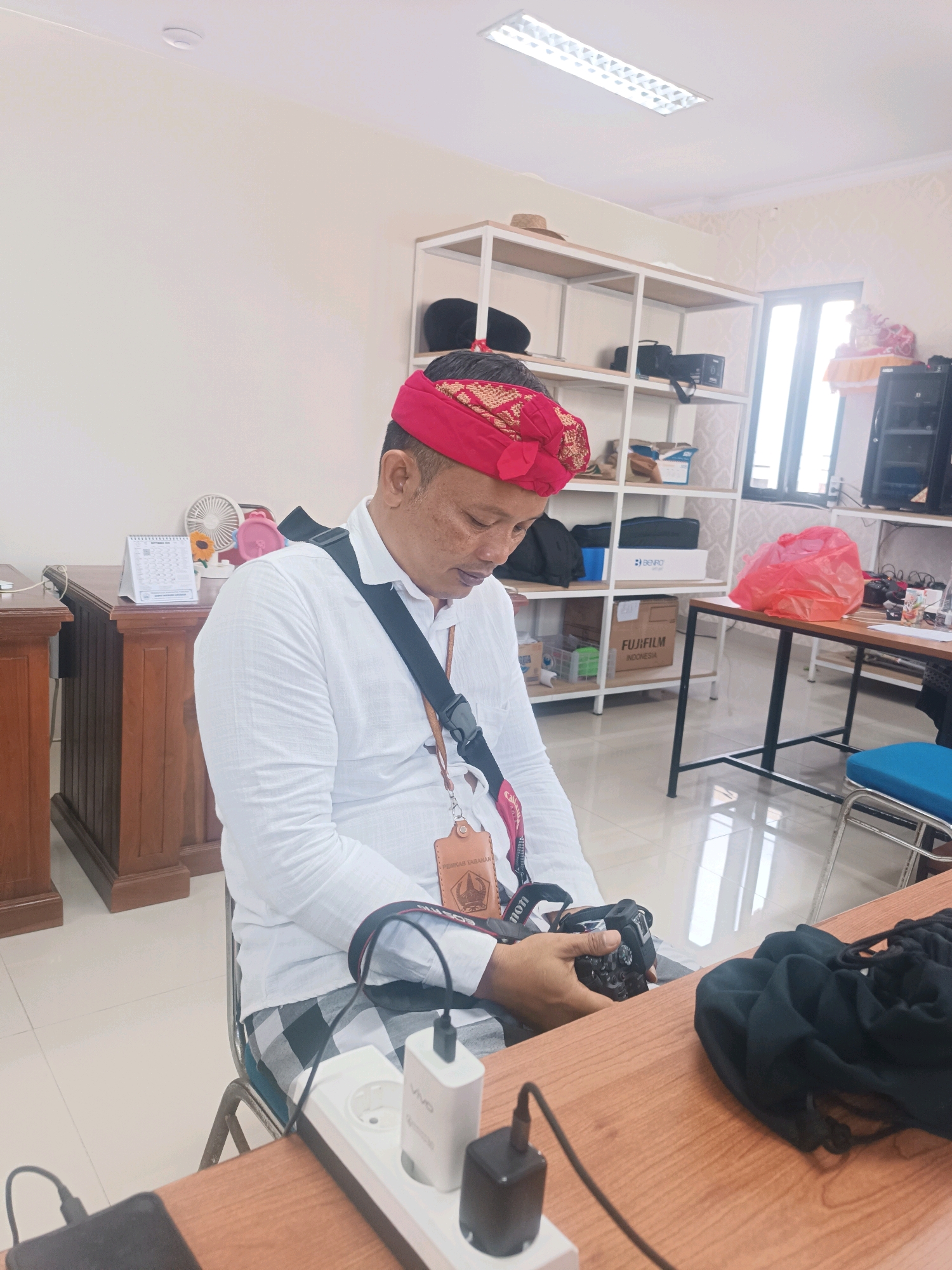 In Tabanan, Indonesia, a man wearing a red traditional headdress and a white long-sleeved shirt sits at a wooden desk, looking down at a black digital camera in his hands. He has a black strap around his neck, from which hangs a tan lanyard with an emblem. He is also wearing a black and white checkered sarong. On the desk in front of him are a power strip with several plugs, a black phone, a black charging cable, and a crumpled black fabric. Behind him, a light-colored wall is visible, with shelves containing various items, including bags, boxes, and electronics. A window is visible to the right, with light streaming through. A wooden desk with drawers is situated to the left of the frame, with a fan and a calendar on it. The overall environment appears to be an office or workroom.