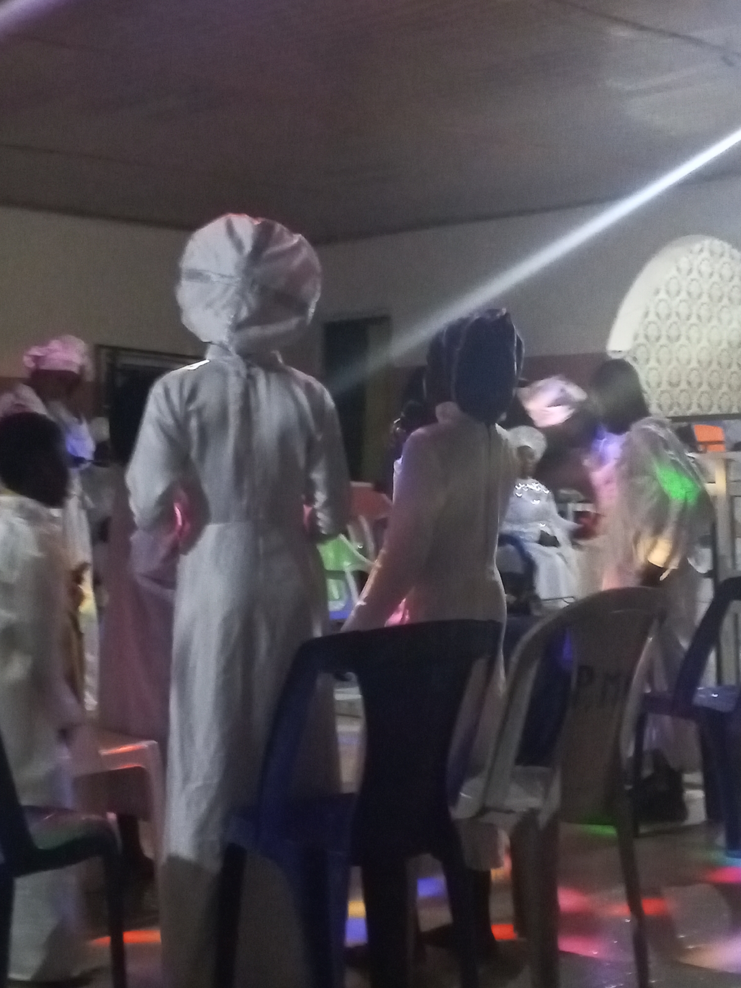 The image depicts a group of people in Lokoja, Nigeria, dressed in white garments and head coverings, possibly attending a religious ceremony or gathering. They are standing and sitting among plastic chairs in an indoor space with patterned wall decorations. The lighting is dim, with a bright light streak across the top of the scene, and colored lights illuminating the floor.
