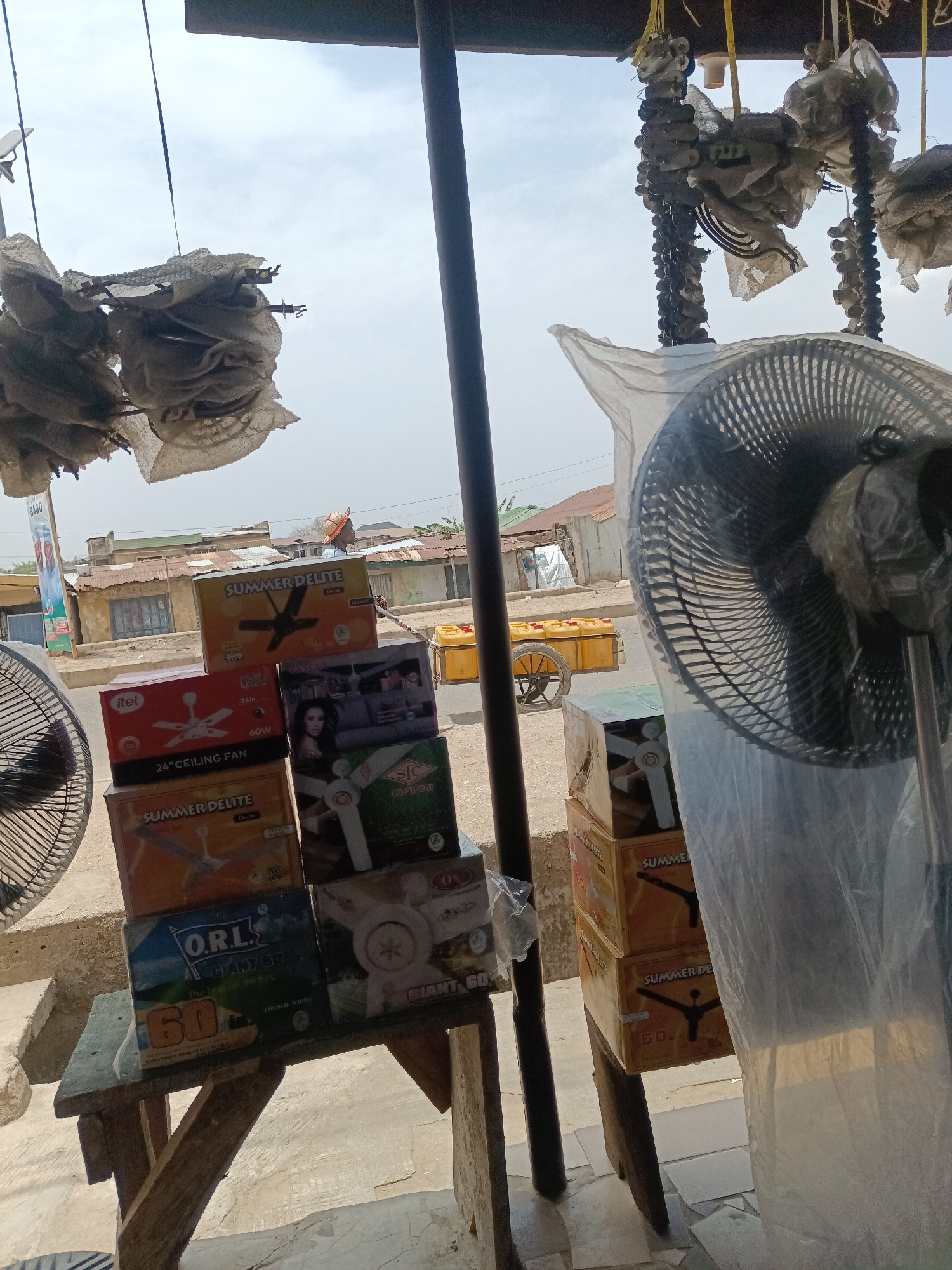 A photograph taken outdoors in Minna, Nigeria, shows a cluttered storefront with various ceiling fan boxes stacked on a wooden table. The brands include "Summer Delite," "Itel," "SIC President," and "O.R.L Giant 60." A standing fan, partially covered in plastic, stands to the right. Outside the storefront, a man wearing a straw hat is walking along the street. A yellow cart filled with yellow containers is visible in the background. The buildings in the background appear to be in a state of disrepair. Hanging from the storefront are what appear to be strings of unidentified objects. The lighting suggests it is daytime.