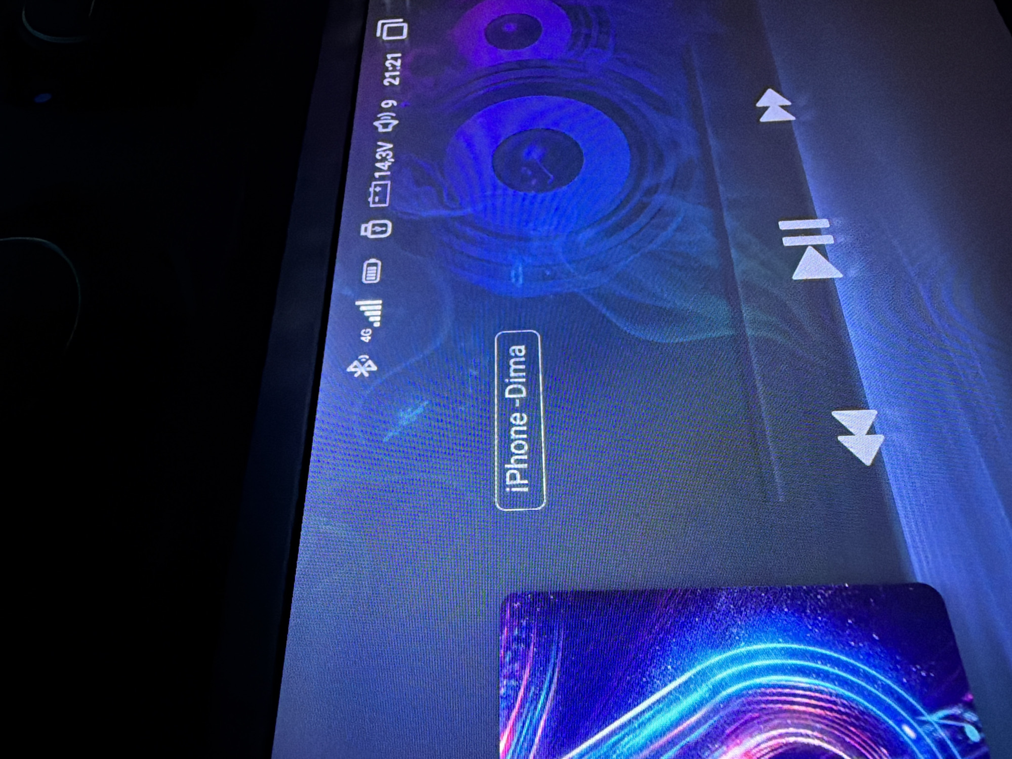 The image displays a close-up of a car's infotainment screen, likely in Pănășești, Moldova, due to the context provided. The screen shows a music player interface. On the left, a colorful, abstract image, possibly album art, is visible. In the center, the text "iPhone - Dima" is displayed, indicating the connected device and user. The top status bar shows icons for Bluetooth, 4G signal strength, battery level, and time (21:21). Navigation buttons for music playback, including rewind, play/pause, and fast forward, are present at the bottom of the screen. The background of the screen features a stylized, circular, blue and purple graphic, resembling sound waves or a speaker cone. No people are visible in the image.