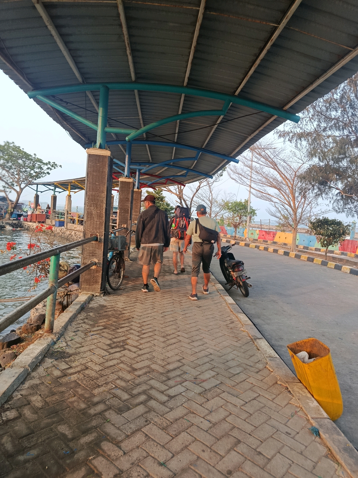 The image depicts a waterfront pathway under a covered structure in Jepara, Indonesia. Three individuals are walking away from the viewer on the paved pathway. The person in the foreground wears a dark jacket, grey shorts, and a baseball cap. Behind them, a person with a backpack wears a light-colored shirt and shorts. To their left, another person with a large backpack is partially visible. A bicycle is parked near a pillar on the left side of the pathway. Adjacent to the pathway is a body of water with some red flowers visible on a railing. Further down the pathway, a motorcycle is parked next to a bright yellow trash bin. The background shows trees and a colorful barrier along the road. The scene appears to be a public area, possibly a pier or promenade, with elements of local commerce visible near the water's edge.