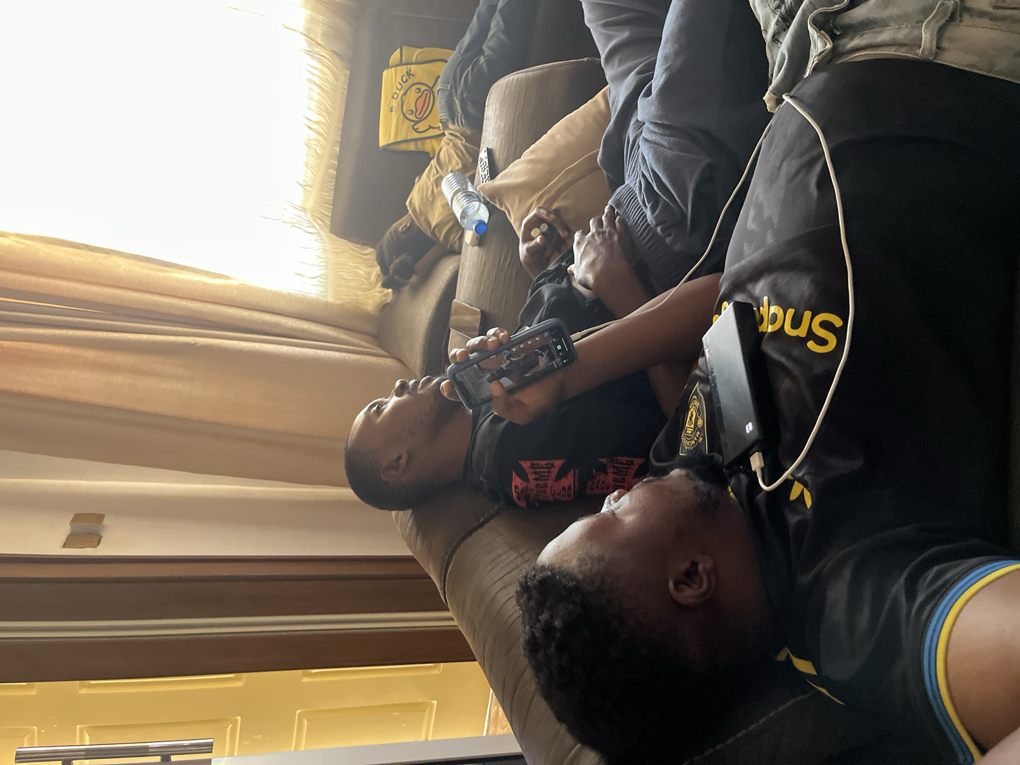 Four individuals are situated on a brown sofa. One individual is viewing a mobile phone. A water bottle is located near another individual. The environment appears to be indoors with curtains visible. The location is Jos, Nigeria.