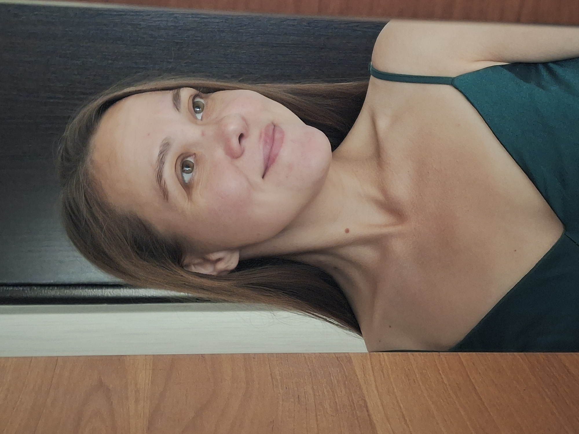 The image shows a close-up of a woman's face and upper torso, taken from a slightly elevated angle. She has light brown hair, which is styled down and frames her face. Her eyes are open and looking upwards, with a slight smile on her lips. She is wearing a dark green spaghetti strap dress, with one strap visible over her left shoulder. The background is divided horizontally. The upper portion is dark and textured, possibly a wall or furniture. The lower portion is a wooden surface, likely a table or floor, with a visible grain. The lighting suggests an indoor setting, possibly during the daytime given the soft, natural light illuminating her face. There are no visible signs of specific activity or event, and the overall mood conveyed is relaxed and contemplative. No text is visible in the image. The location context of Chaikovsky, Russia is not visually apparent from the image itself.
