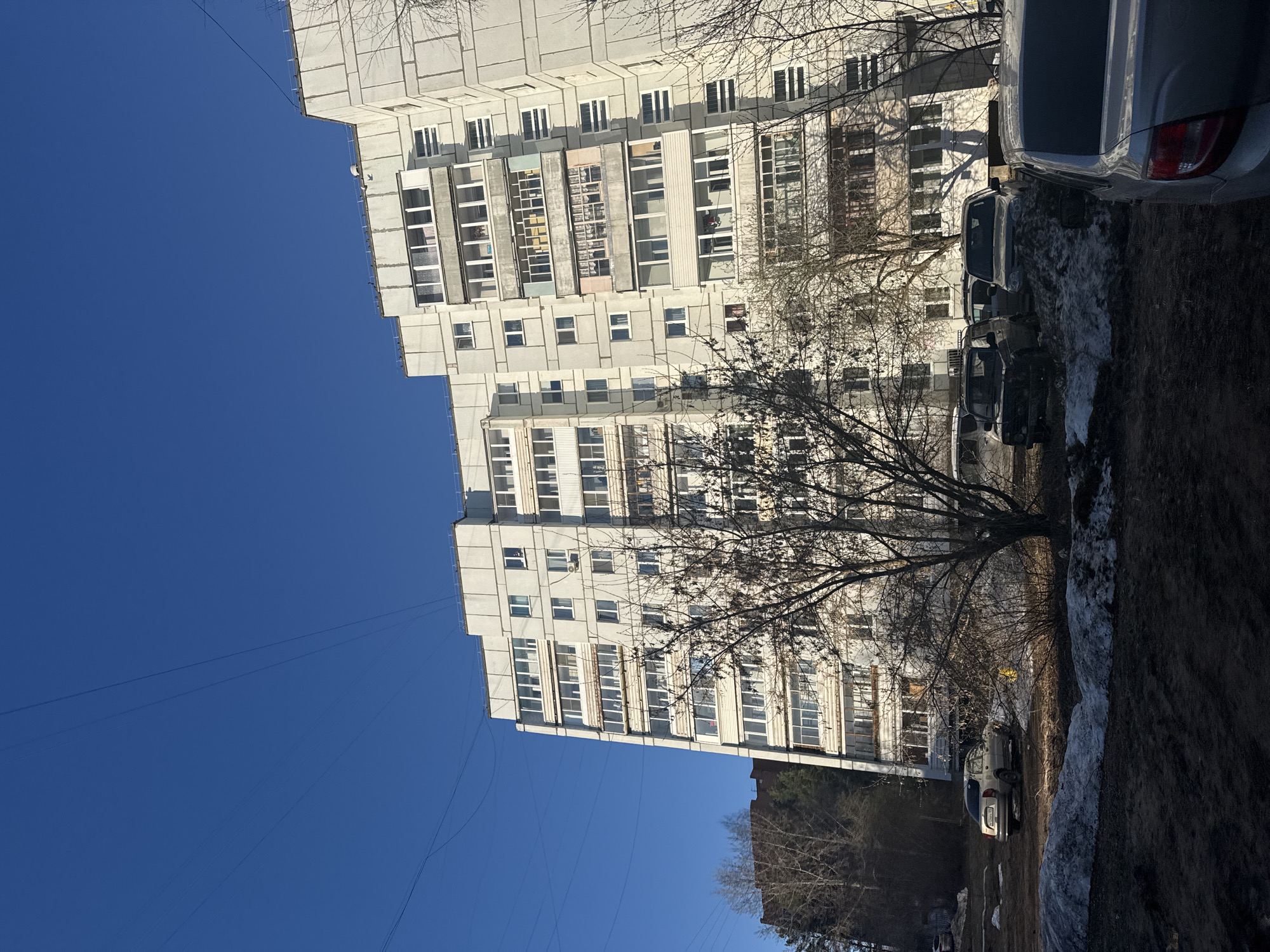 The image depicts a clear blue sky above a Soviet-era apartment building in Chaikovsky, Russia. The building is multi-storied and constructed of concrete panels with rows of rectangular windows. The windows are a mix of clear glass and some are covered with curtains or blinds. The building is partly obscured by bare tree branches in the middle ground. Below the building, the ground shows patches of snow mixed with dark earth. Several parked cars are visible in front of the building. Electrical wires are strung across the sky near the upper left corner of the frame.