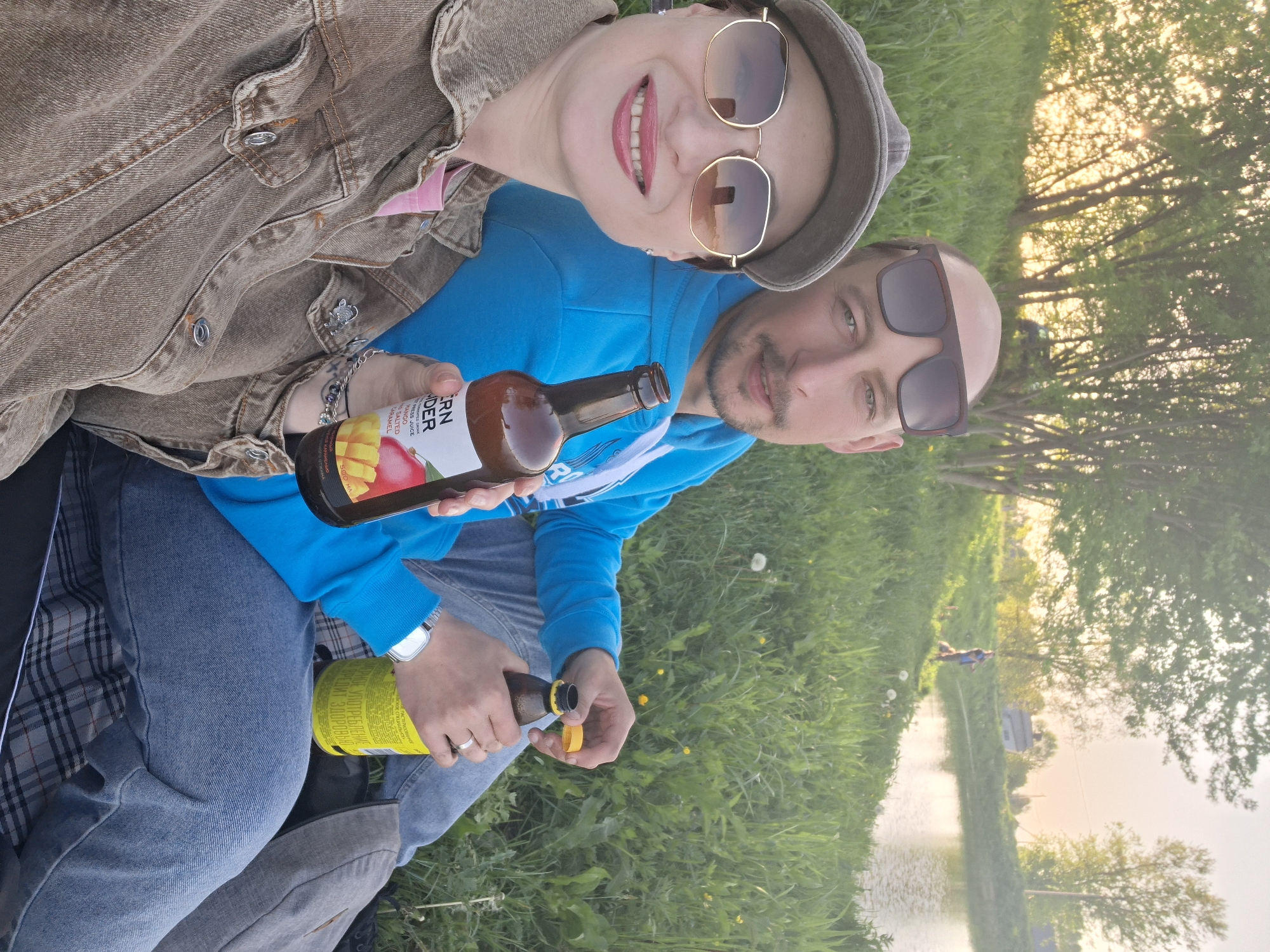 Here is a matter-of-fact description of the image:
A couple is taking a selfie outdoors, sitting in the grass near a body of water, possibly a lake or river. The woman is wearing a brown denim jacket, sunglasses, and a hat. The man is wearing a blue hooded sweatshirt and sunglasses. They are holding bottles of beer or cider. The environment includes trees, green grass, and a body of water that reflects light, possibly sunset. The text on one of the bottles suggests it's a mango and salted caramel flavored drink. The location may be in Забалоцкі сельскі Савет, Belarus, based on text in the bottle.