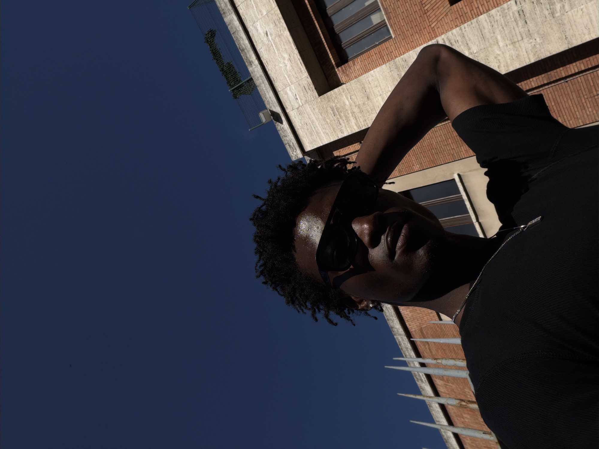 This is a low-angle, Dutch tilt shot of a person wearing sunglasses and a black, high-necked shirt. The person has dark skin and curly hair. Their right arm is raised and resting on their head. A silver chain is visible around their neck. The background consists of a building with brick and concrete elements, and a clear blue sky. The sunlight is strong, casting shadows and highlighting the person's face and the building's texture. The image suggests it is daytime. The building's architecture, particularly the brickwork and the windows, and the fence in the foreground with its pointed metal spikes, hint at an urban environment, possibly in Milan, Italy. There is no visible text in the image.