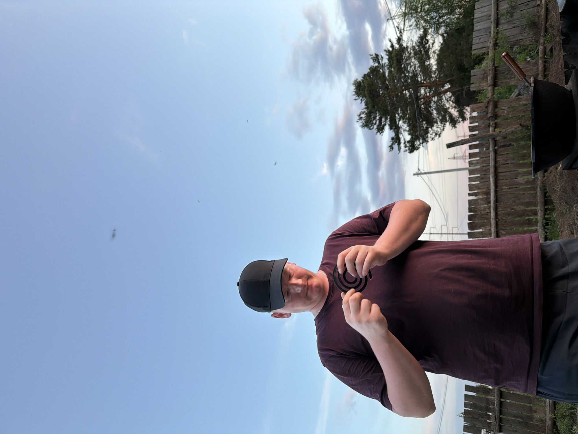 A man in a black baseball cap and a burgundy t-shirt is shown from the waist up. He is standing outdoors, holding a mosquito coil. The sky is blue with scattered clouds. In the background are trees, a wooden fence, and power lines. The location may be Chaikovsky, Russia.