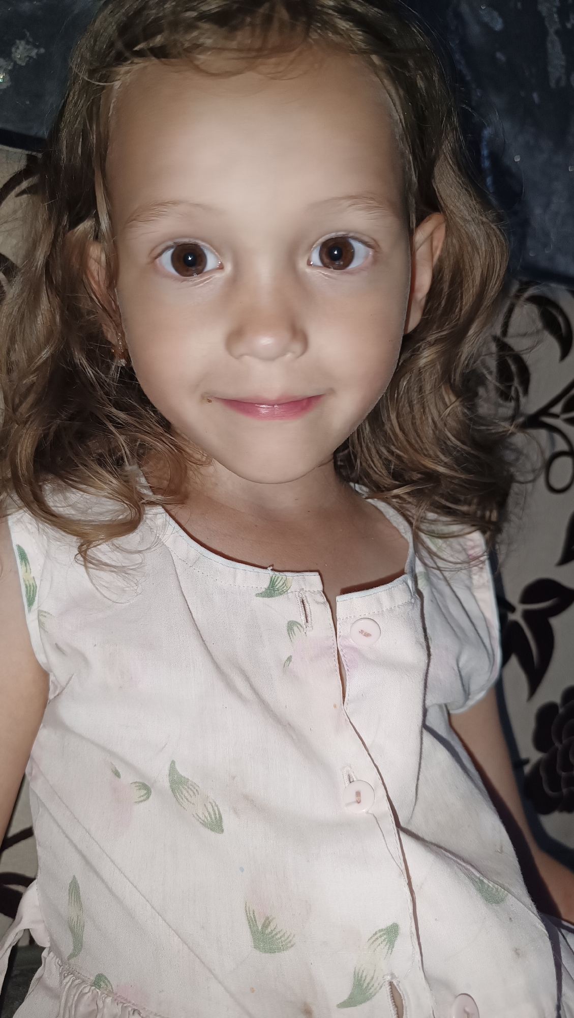 A close-up portrait shows a young girl with light brown curly hair and brown eyes looking directly at the camera. She is wearing a light pink dress with a floral pattern. The background is out of focus and appears to be indoors, with a patterned surface visible behind her. The lighting suggests an indoor setting, possibly a home environment. There is no information in the image that indicates a location in the city of Karsha, Russia.