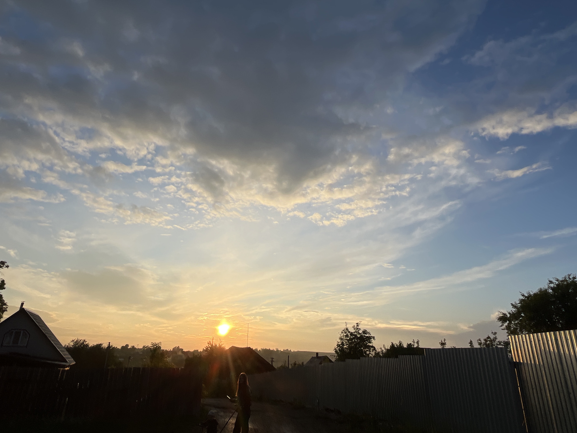The image depicts a rural scene at dusk in Fokino, Russia. The sky is a mixture of blue and gray clouds, with the sun low on the horizon, casting a warm, orange glow. In the foreground, a dirt path is visible, with a silhouette of a person walking away from the camera, accompanied by a smaller figure, possibly a child or a pet. To the left, a dark silhouette of a house with a distinctive pointed roof and a window is seen. A dark fence runs along the left side of the path. To the right, a tall, corrugated metal fence dominates the mid-ground. Trees and other structures are faintly visible in the background, silhouetted against the sunset. The overall impression is one of a quiet, rural evening.