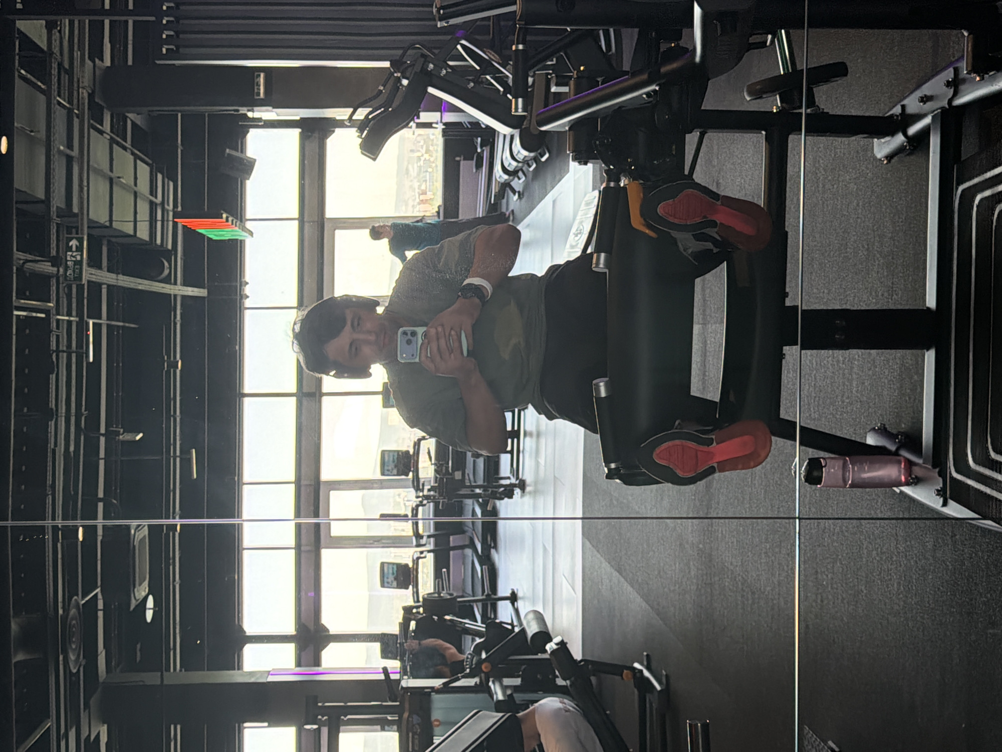 The image captures a selfie taken in a gym mirror, showing a young man seated on a leg press machine. He is wearing a dark green t-shirt, dark shorts or pants, a white wristband, and a watch on his left arm, while holding a smartphone with a light blue case to take the photo. He has dark hair and is looking directly at the phone.
The gym features large windows in the background, revealing a bright daytime view of an urban or suburban landscape with distant buildings and what appear to be hills or mountains. The gym interior is modern, with dark walls and flooring, and various fitness machines are visible reflected in the mirror. Another person can be seen further back in the gym, near the windows.
Notable details include the man's red-soled athletic shoes on the leg press footplate and the clear reflection distorting depth. On a dark column to the left, a green and red light strip is visible, along with a white "EXIT" sign. Below "EXIT", the Cyrillic word "ВЫХОД" (Vykhod, meaning "EXIT") is also visible, consistent with the location context of Krasnoyarsk, Russia. The overall lighting suggests it is daytime, with ample natural light entering the facility.