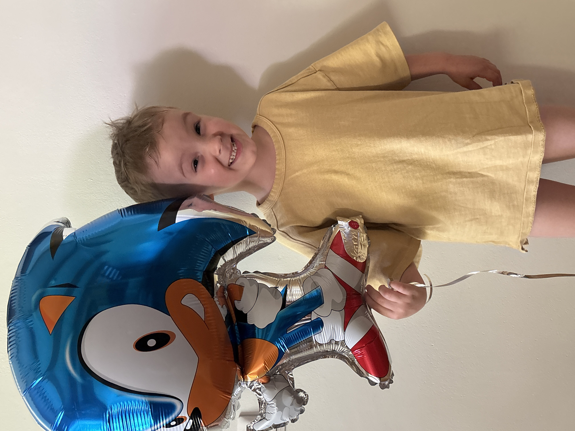 A young child smiles while holding a Sonic the Hedgehog balloon indoors. The balloon is comprised of a blue, metallic depiction of Sonic's head and a red and white representation of his shoe. The child wears a short-sleeved yellow shirt and appears to be standing, with the lower portion of their body partially visible. The background is a plain, light-colored wall. There is no indication of the city of Chaikovsky, Russia, within the image.