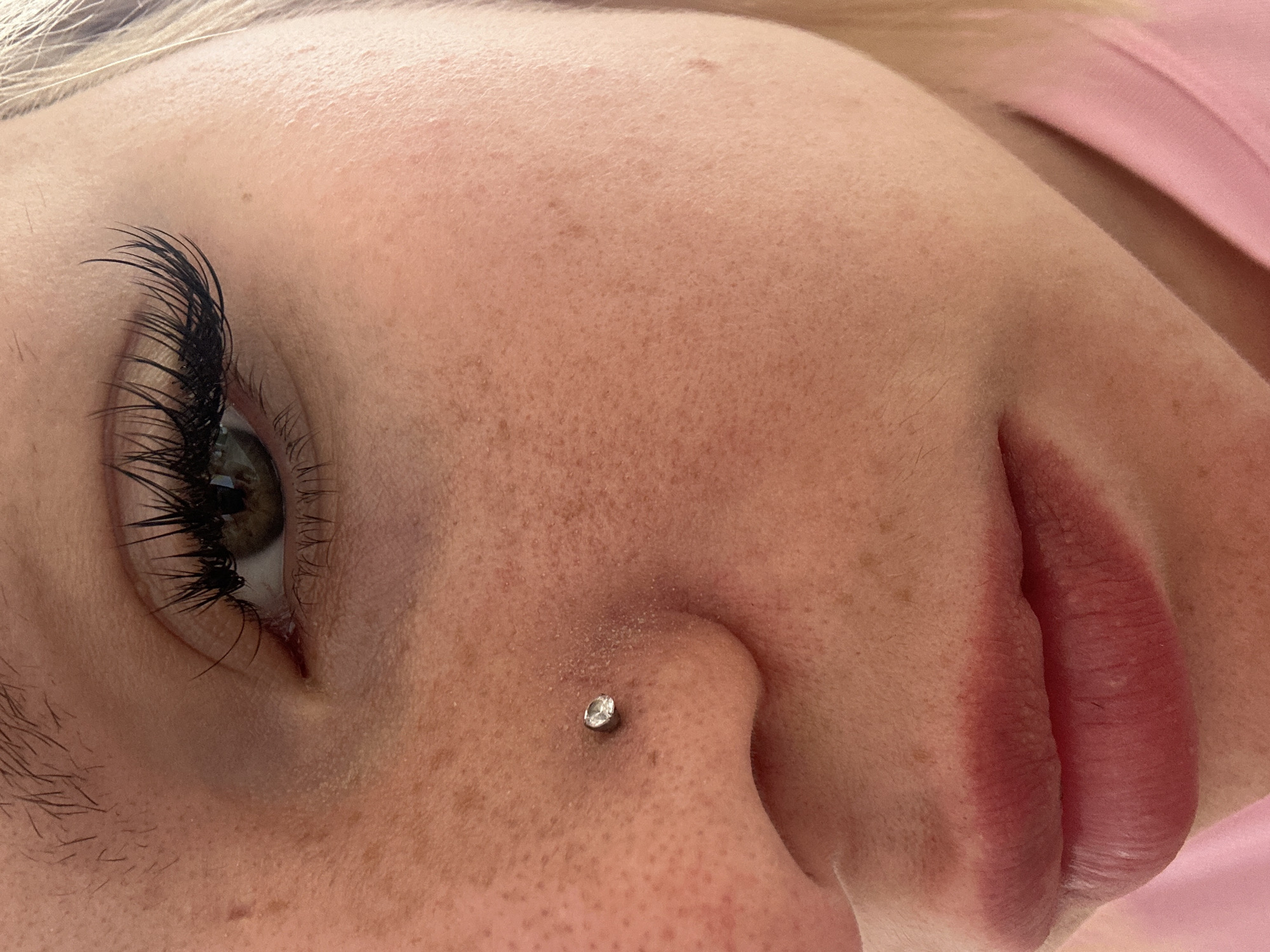 A close-up, eye-level shot focuses on the right side of a person's face, from the eye down to the lips. The person has light skin with freckles and appears to be wearing full eyelash extensions. A small, silver stud with a clear gem is visible in the person's nostril. The background is blurred, with a hint of pink fabric on the lower right. The image appears to be taken indoors, possibly in a residential setting. There is no indication of the city of Budva, Montenegro.