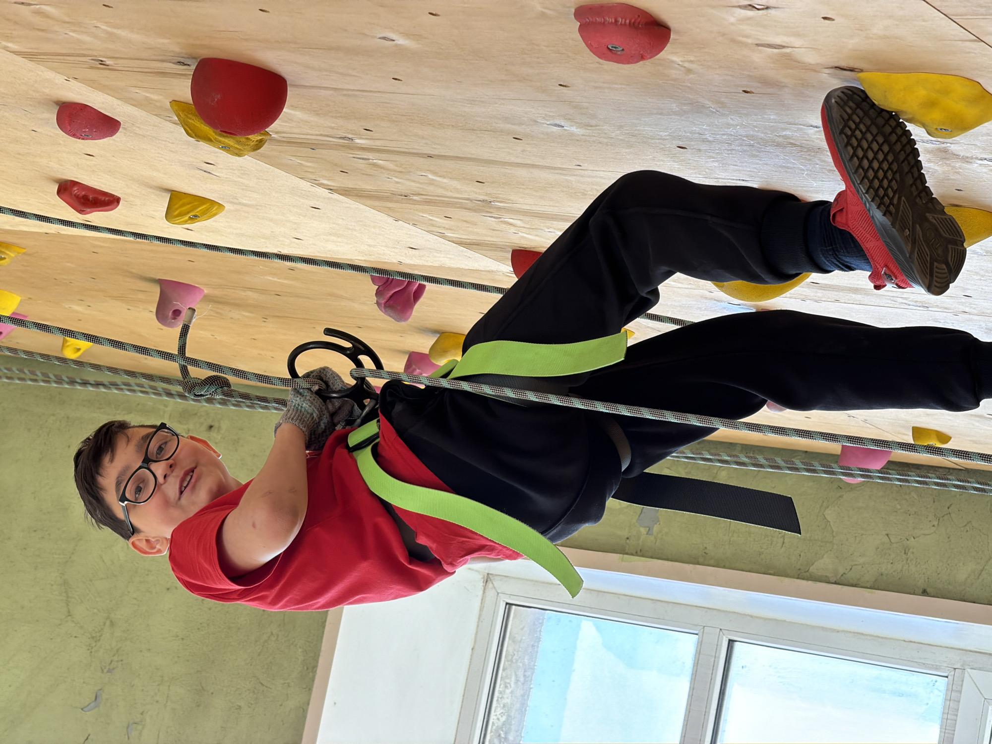 This image depicts a young boy engaged in rock climbing at an indoor facility, likely in Izhevsk, Russia. The boy, appearing to be in mid-climb or resting, is suspended against a wooden climbing wall, with the image rotated such that he appears horizontal. He wears black-framed glasses, a red t-shirt, black pants, and red athletic shoes. His climbing gear includes a harness with bright neon green straps and grey fingerless gloves, and he is secured by a climbing rope.
The climbing wall, made of light-colored wooden panels, is adorned with numerous colorful holds in shades of red, yellow, and pink, suggesting various routes. The boy looks directly at the camera with a slight smile, conveying engagement and enjoyment. His right foot is placed on a yellow hold, and his hands are gripping the rope.
In the background, a large window provides natural light, revealing a bright, possibly overcast, sky. The wall below the climbing structure is painted a light green, showing some texture. The overall scene captures an active and fun sporting environment. No visible text is present in the image.