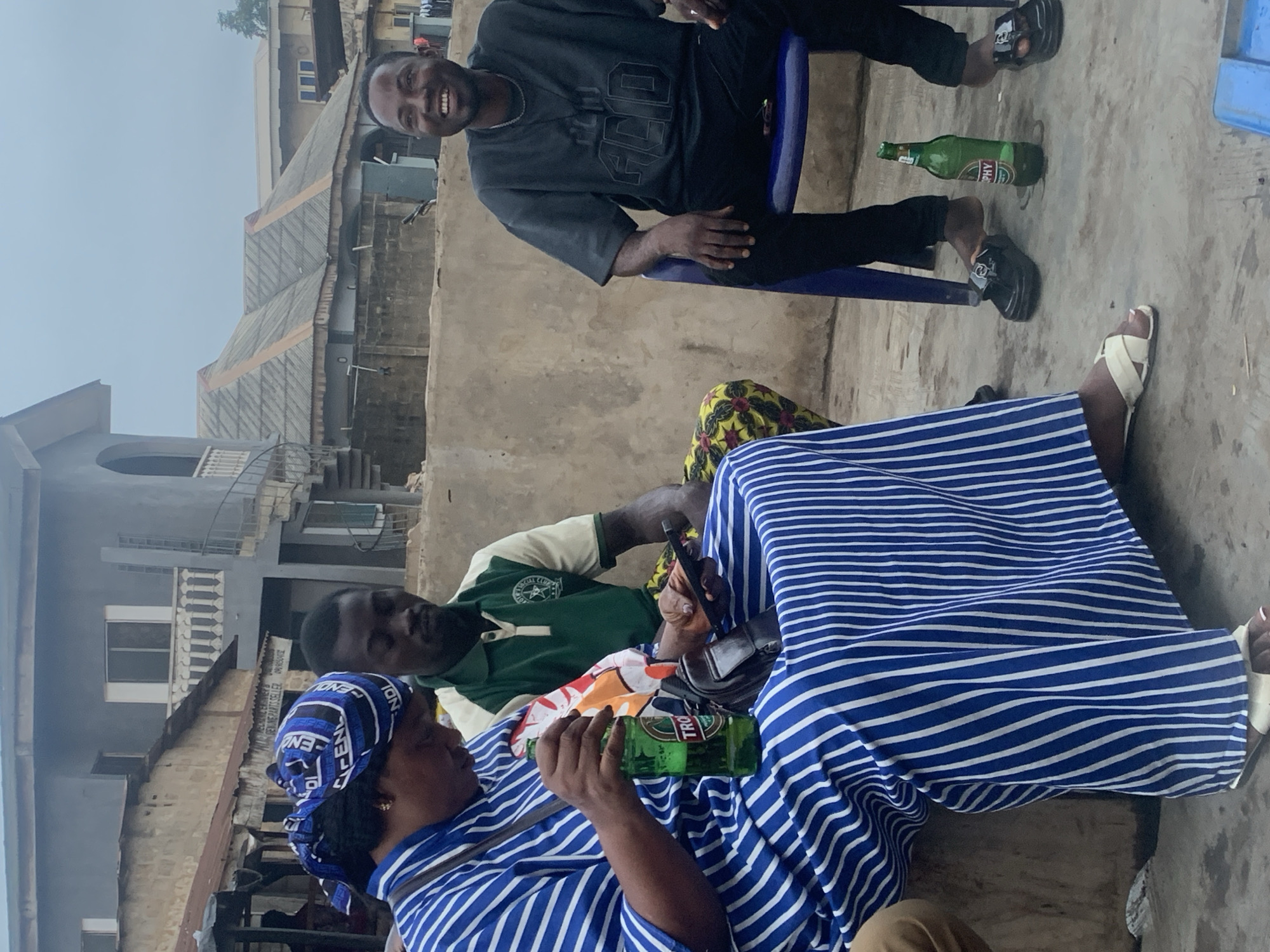 The image depicts an outdoor scene in Shagamu, Nigeria, featuring three individuals. In the foreground, a woman wearing a blue and white striped dress and a blue headscarf is seated, holding a bottle of Trophy beer and a mobile phone. To her left, a man in a green and white polo shirt is also seated. To the right, another man in a dark gray t-shirt and black pants is seated on a blue chair, smiling. Behind the people, there are buildings with concrete walls and corrugated iron roofs, suggesting an urban or semi-urban environment. A second bottle of Trophy beer is visible on the ground near the man in the gray t-shirt. The ground is paved with concrete. The overall lighting suggests an overcast day.