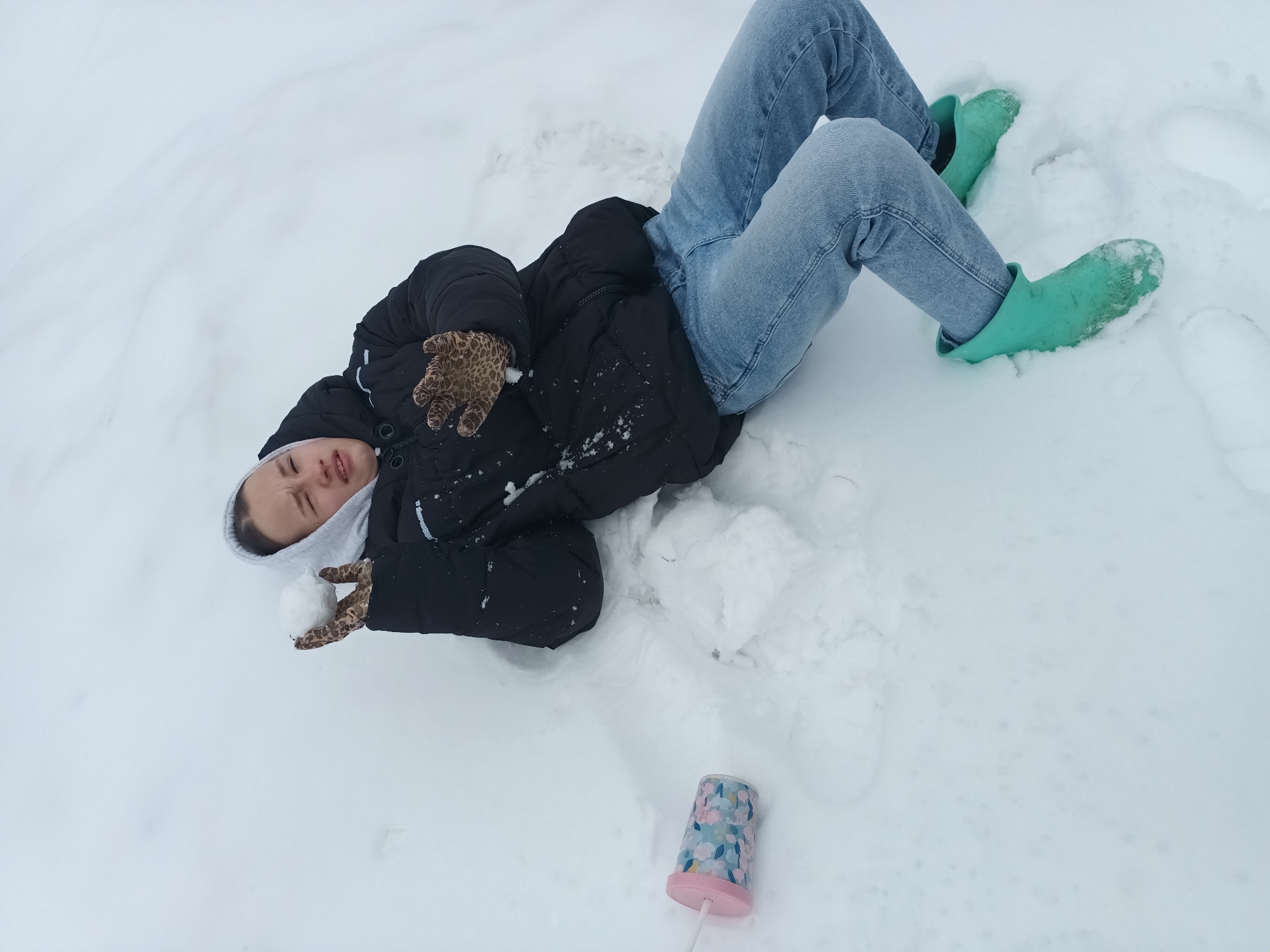 A person is lying in the snow. The person is wearing a black puffy coat, light blue jeans, and bright green boots. The person is also wearing patterned gloves and holding a snowball in one hand. A cup with a straw is lying on the snow near the person. The location is Ольховка, Russia. The environment is snowy.