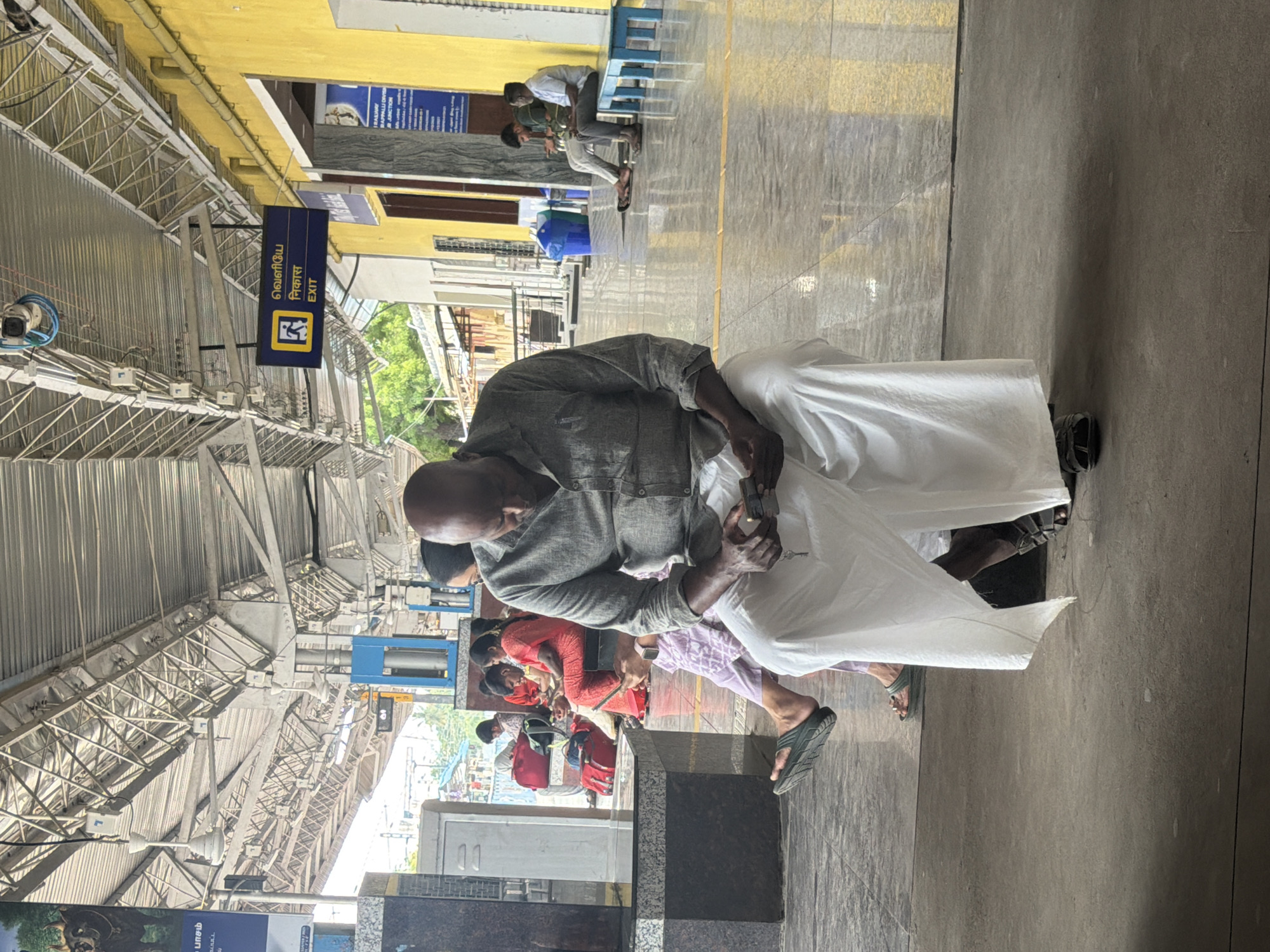 The image depicts a man sitting at a train station in Thanjavur, India. He is bald, wearing a grey shirt and white dhoti, and appears to be looking at a mobile phone in his hands. Several other people are visible in the background, some sitting on benches, and others standing. The environment is an open-air platform with a covered roof supported by metal beams. There are yellow and blue signs on the wall, one of which indicates "EXIT" in multiple languages. The ground is tiled, and there are some potted plants visible in the distance. The overall scene suggests a typical day at a busy Indian railway station.