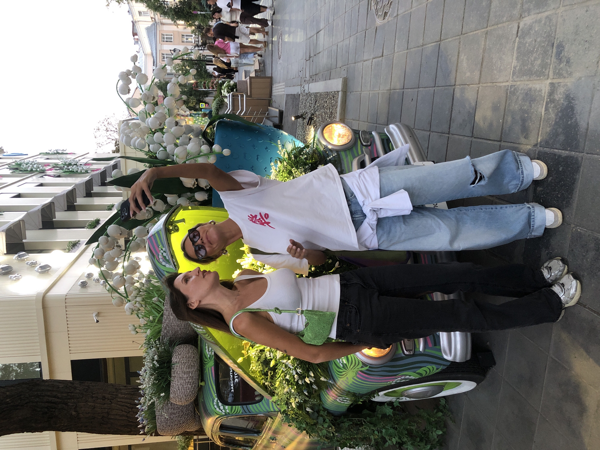 Two women are taking a selfie in front of a vintage car decorated with lilies of the valley. The car is green with colorful wavy stripes and has its hood open, revealing lush greenery and more flowers. The woman on the left is wearing a white tank top, black jeans, and silver sneakers. She has a green beaded handbag. The woman on the right is wearing a white t-shirt with pink lettering, light blue distressed jeans, and white sneakers. She has sunglasses on and is holding her phone up to take the photo. They are standing on a paved sidewalk. In the background, there are people walking, buildings, and trees. The scene appears to be on a city street, possibly in a pedestrian area, given the festive decoration and the presence of onlookers. The car's license plate is partially visible and appears to be from Russia. The overall atmosphere is cheerful and celebratory, possibly during an event or a street festival. This image is likely from a public space in Moscow, Russia, as indicated by the Russian text on the car and the architecture visible in the background.