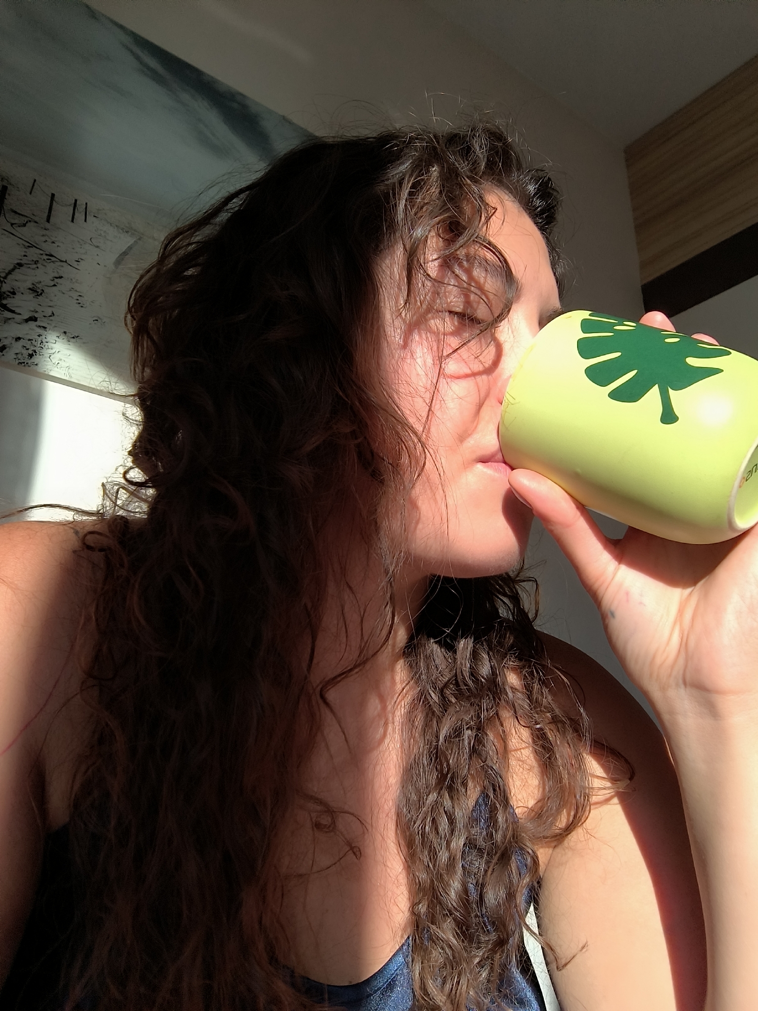 A woman with long, dark curly hair is drinking from a yellow mug decorated with a green monstera leaf. She is holding the mug with her right hand and it is partially obscuring her face. Sunlight is falling on her face, hair, and arm. She is wearing a blue top. In the background is an interior, including an artwork, possibly a painting of a coastal scene, hanging on the wall. The photograph was taken indoors, likely near a window allowing sunlight to enter. There is no information about the location being in Budva, Montenegro.