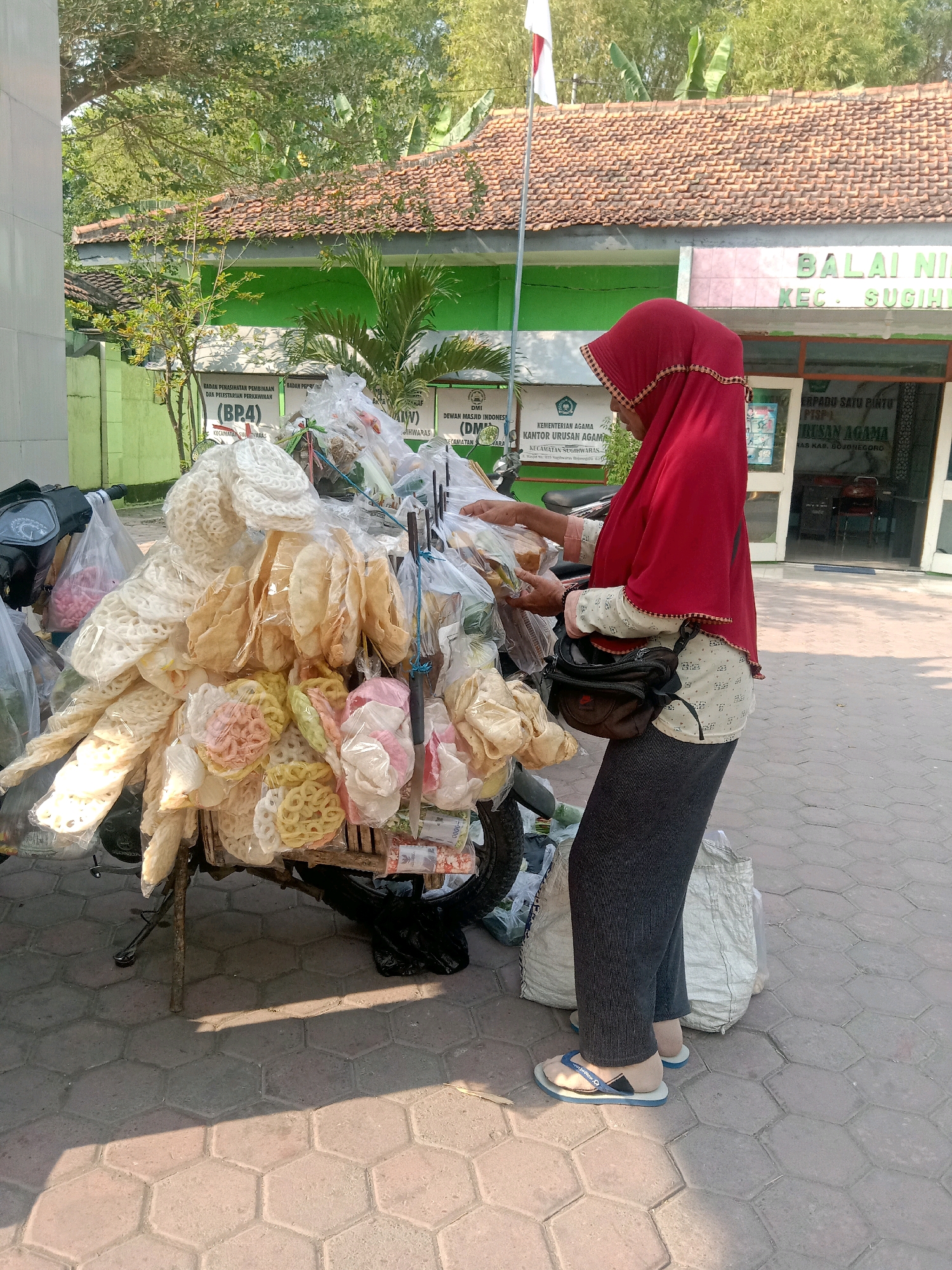 A woman in a red hijab and a patterned blouse stands beside a cart laden with packaged snacks, likely crackers, in Sugihwaras, Indonesia. The cart is attached to a motorcycle. The woman is reaching into the cart, holding a snack. In the background, a building with a sign reading "BALAI NIA KEC. SUGIHWARAS" is visible, suggesting the location is a sub-district office. The ground is paved with hexagonal tiles. The scene is lit by sunlight, casting shadows.