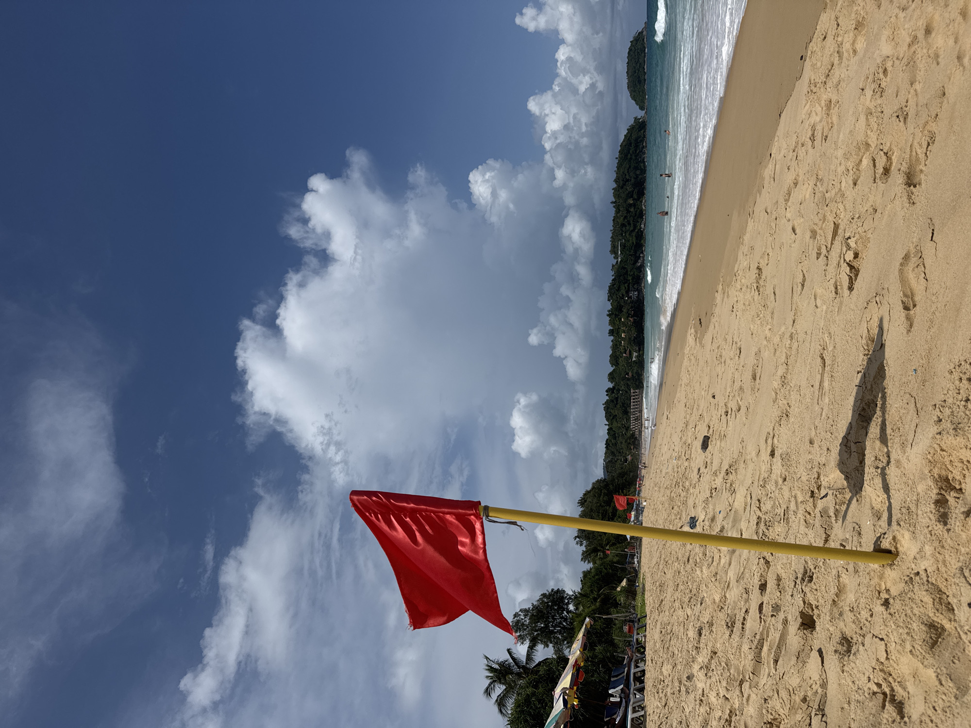 A red flag flies from a yellow pole in the foreground of a sandy beach. The beach is lined with umbrellas and lounge chairs, and the ocean with waves is visible in the background. Lush green hills with buildings are visible in the distance. The sky is blue with white clouds. This is Karon Beach, Thailand.