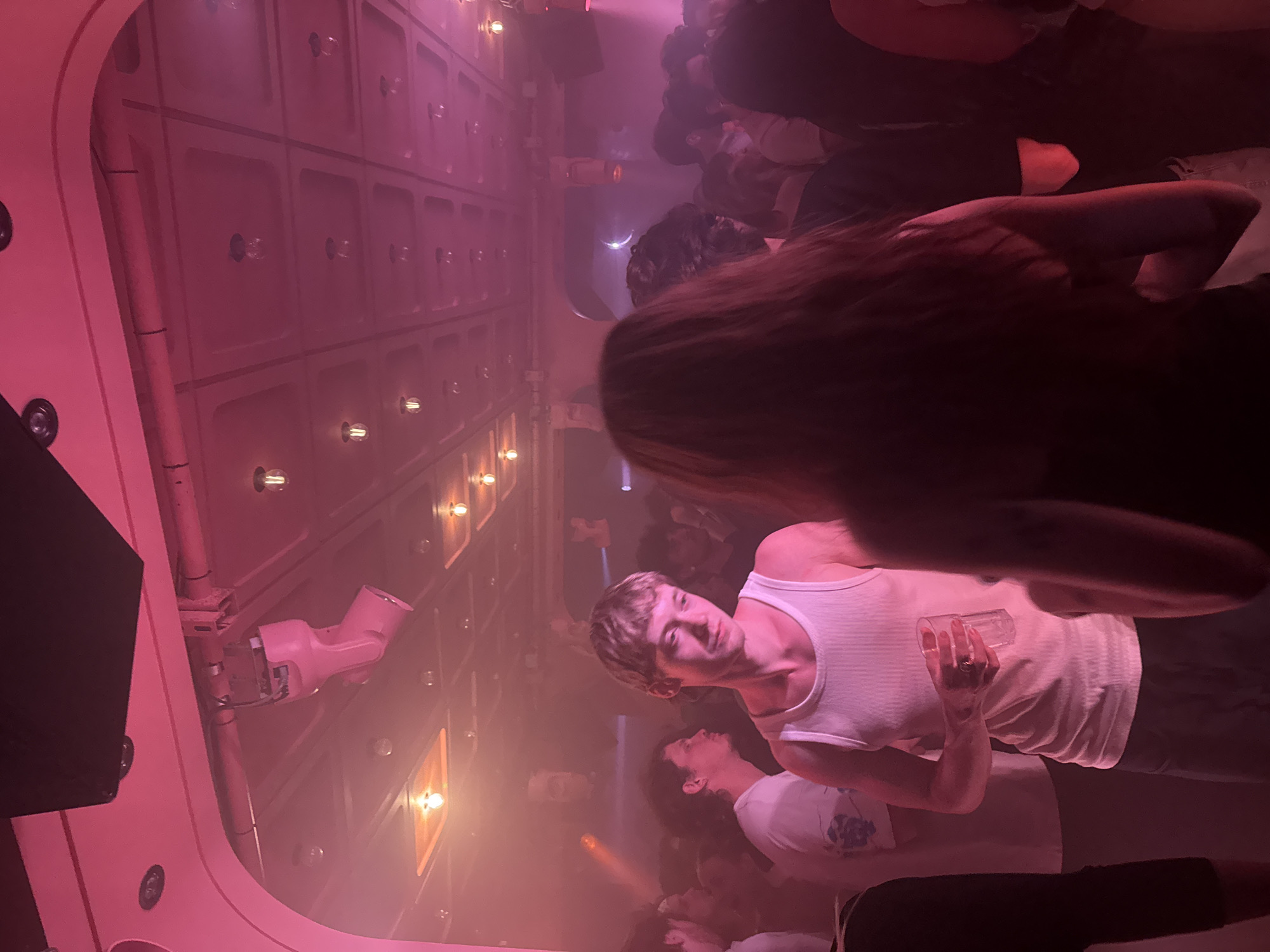 The scene is indoors, illuminated in pink light. The location may be a bar or club, given the crowd of people. A person in a white tank top holds a glass. The walls have a grid-like pattern with light fixtures. Overhead, there's a pink structure with ventilation or piping. The city of Paris cannot be confirmed from this image alone.