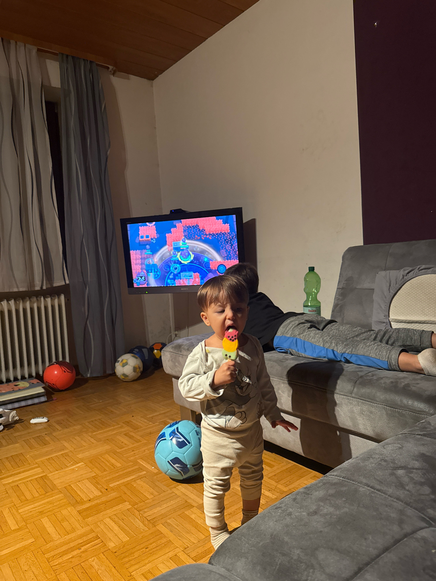 A young child, appearing to be about 2-3 years old, stands in the foreground of a dimly lit living room. The child is wearing a light-colored long-sleeved shirt with a cartoon character design and ribbed beige pants. They are holding a colorful popsicle to their mouth and looking towards the viewer. To the left of the child, a television displays a vibrant video game with animated characters. Several soccer balls are scattered on the wooden floor, including a blue and white one in the mid-ground and a red one to the left, near a stack of books. A grey sectional sofa is prominent in the background, with a second, older child or adult lying on it, partially obscured by the younger child. A green plastic bottle sits on the sofa. The room has striped curtains covering a window on the left and a dark purple wall section on the right. The overall impression is of a casual, indoor family setting, likely a home in Überherrn, Germany.