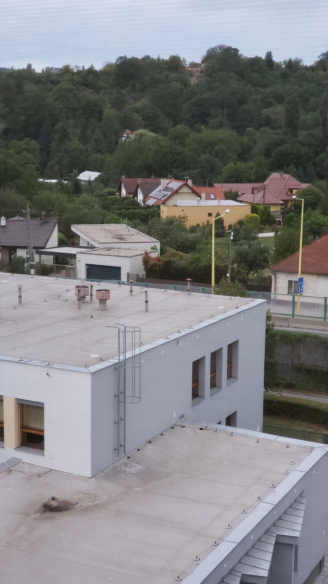 The image shows an elevated view of a residential area in Prešov, Slovakia, with a focus on the rooftops of several buildings. In the foreground, the flat, light-colored roof of a modern, grey, multi-story building is visible, featuring several ventilation pipes and a metal ladder attached to its side. Another, lower building with a similar flat roof is partially visible in the lower right corner.
Beyond this, a cluster of houses with pitched roofs of various colors, including red tiles and grey shingles, are nestled amongst dense, dark green trees that cover a hillside. Some of these houses have solar panels on their roofs. A yellow lamppost stands prominently in the middle ground. The background consists of a heavily wooded hillside with faint signs of human habitation, possibly a few more houses or structures obscured by foliage. The sky is overcast, suggesting a cloudy or a subdued lighting condition. No people are visible in the image.