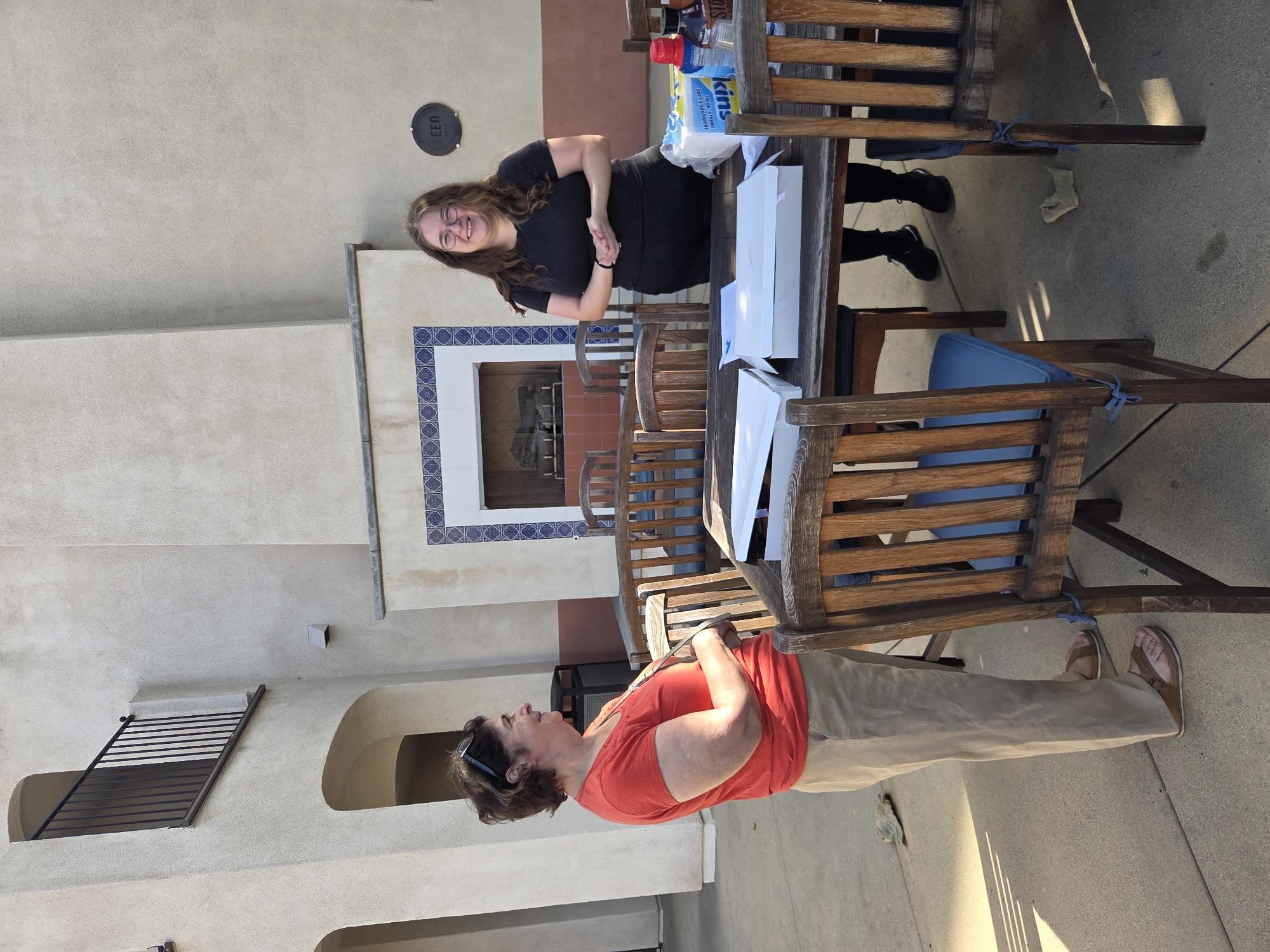 Two women are standing outdoors in what appears to be a patio area. The woman on the left, wearing a red t-shirt and tan pants, is standing with her arms crossed and looking to the left. The woman on the right, wearing a black t-shirt and black pants, is standing with her hands clasped in front of her and smiling at the camera. They are standing behind a wooden table, on which there are several white boxes, a package of paper towels, and some bottles. In the background, there is a building with a fireplace. The scene is lit by natural light, casting shadows on the ground. The location appears to be a residential or communal area, possibly in Glendale, California, given the architectural style and vegetation, although this cannot be confirmed from the image alone.