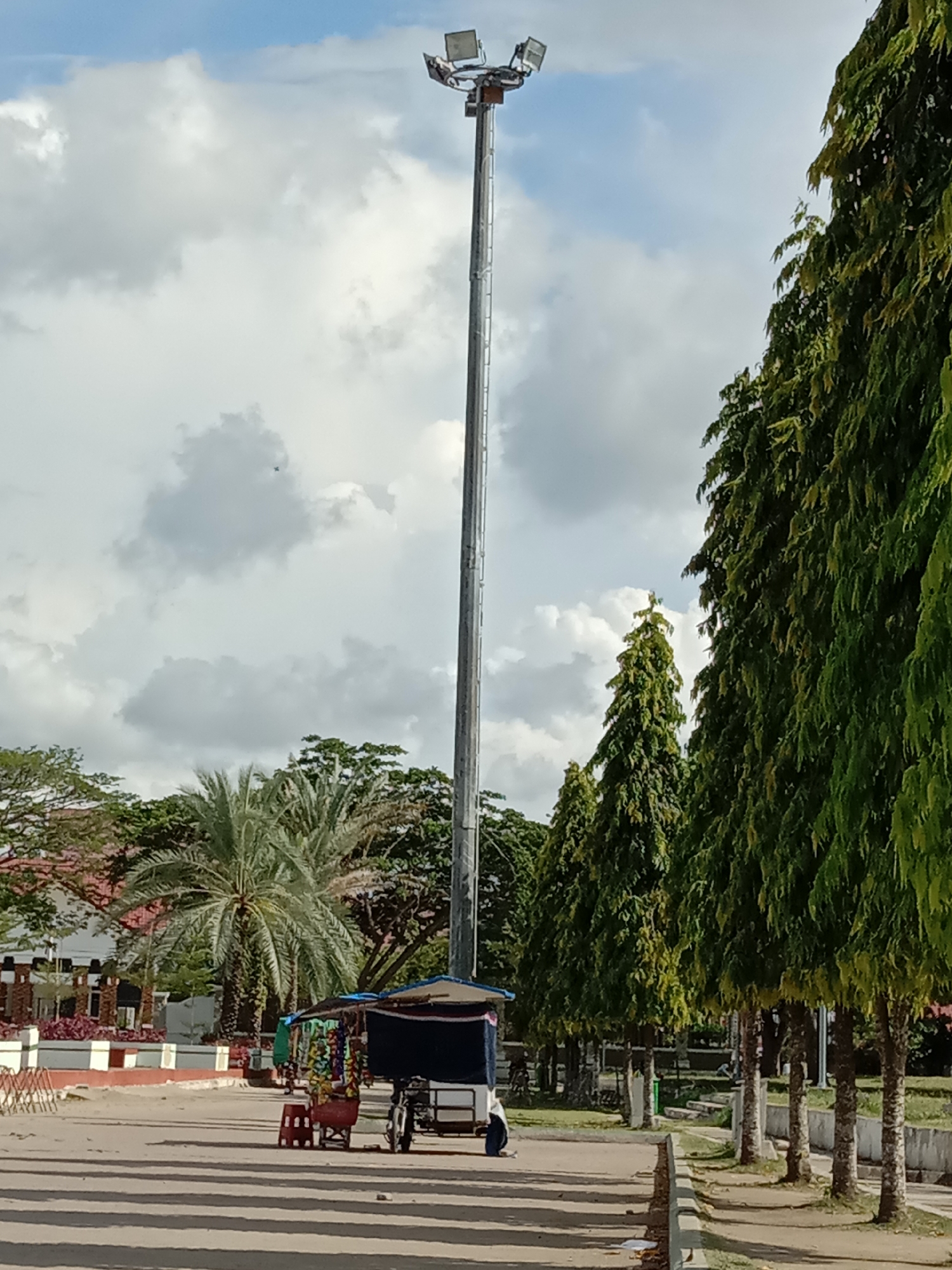 The photograph shows a public space in Lhokseumawe, Indonesia, dominated by a tall metal lighting tower with four directional spotlights at its apex, set against a backdrop of a cloudy sky. A street vendor's cart, selling snacks and beverages, is positioned in the foreground, with a moped parked alongside. Adjacent to the vendor's area, a row of tall, evergreen trees lines the side of the paved walkway. In the distance, a low-rise building with a tiled roof and decorative fencing can be seen. The scene is bathed in daylight, casting distinct shadows across the pavement.