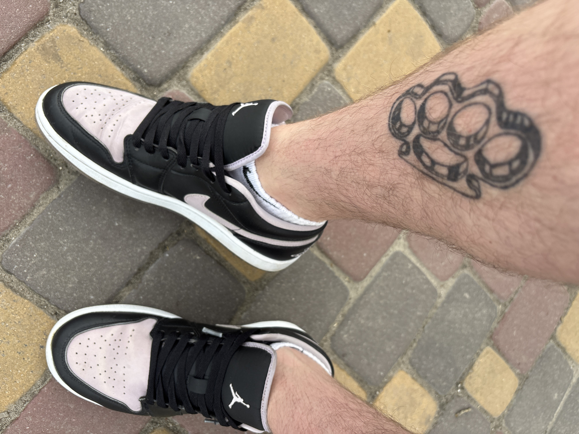 The image shows a close-up, overhead view of a person's legs and feet standing on a sidewalk paved with rectangular bricks of various colors, including gray, yellow, and red. The person has a tattoo of brass knuckles on their left calf. They are wearing a pair of black and light pink Nike Air Jordan 1 sneakers with white socks. The scene appears to be outdoors in a city, likely Dnipro, Ukraine, given the context provided. The lighting suggests it is daytime.