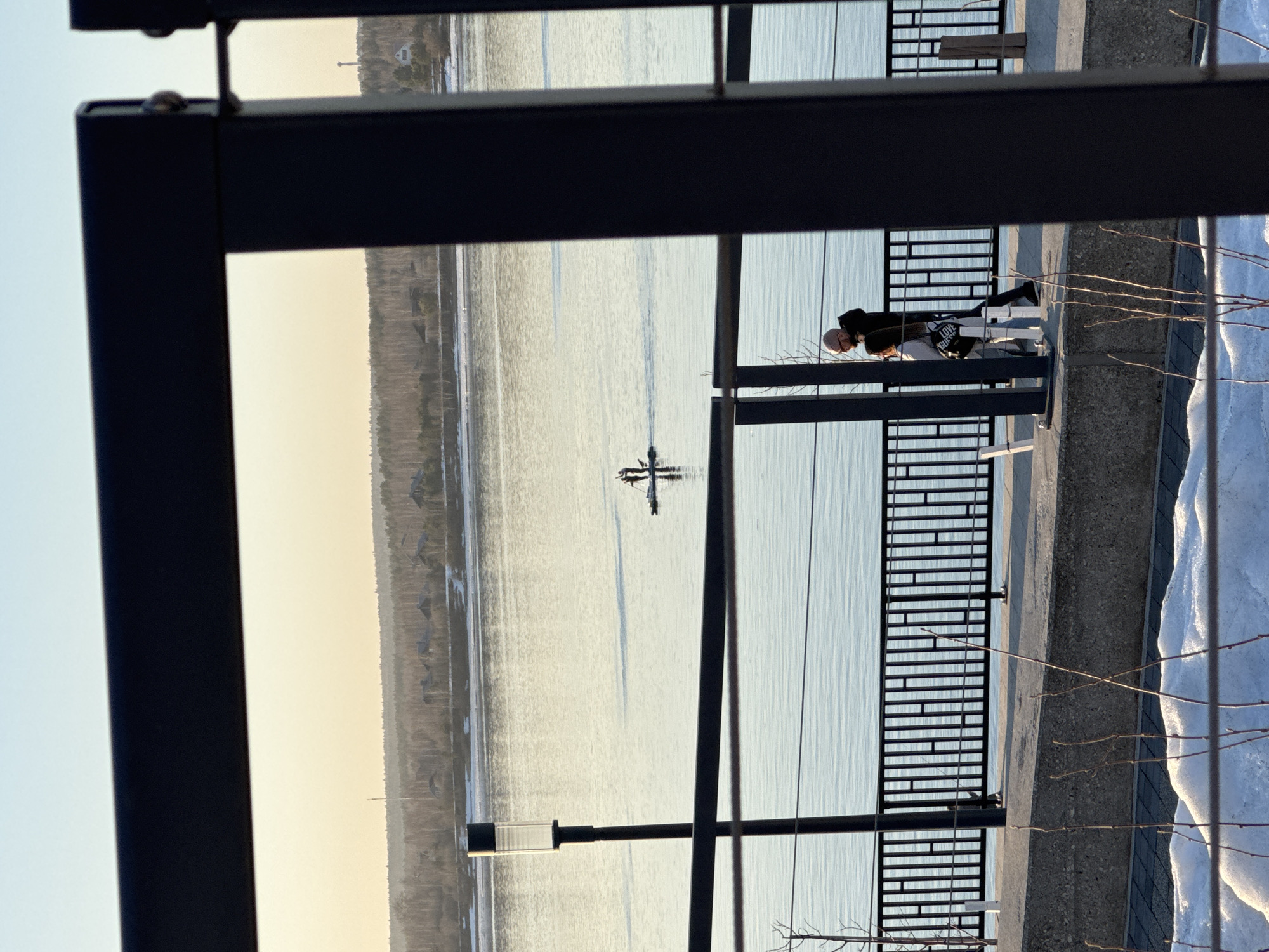 The image depicts a waterfront scene, possibly in Perm, Russia, given the urban development and natural landscape. A couple is visible on a concrete pier or walkway, situated behind a black metal railing. The couple appears to be standing and interacting, possibly taking in the view. Beyond the railing is a body of water, presumably the Kama River, with a distinctive river transport landmark. The opposite bank features a treeline and buildings. There is snow along the edge of the walkway. The image is taken through a partially obstructed view of a dark metal frame.