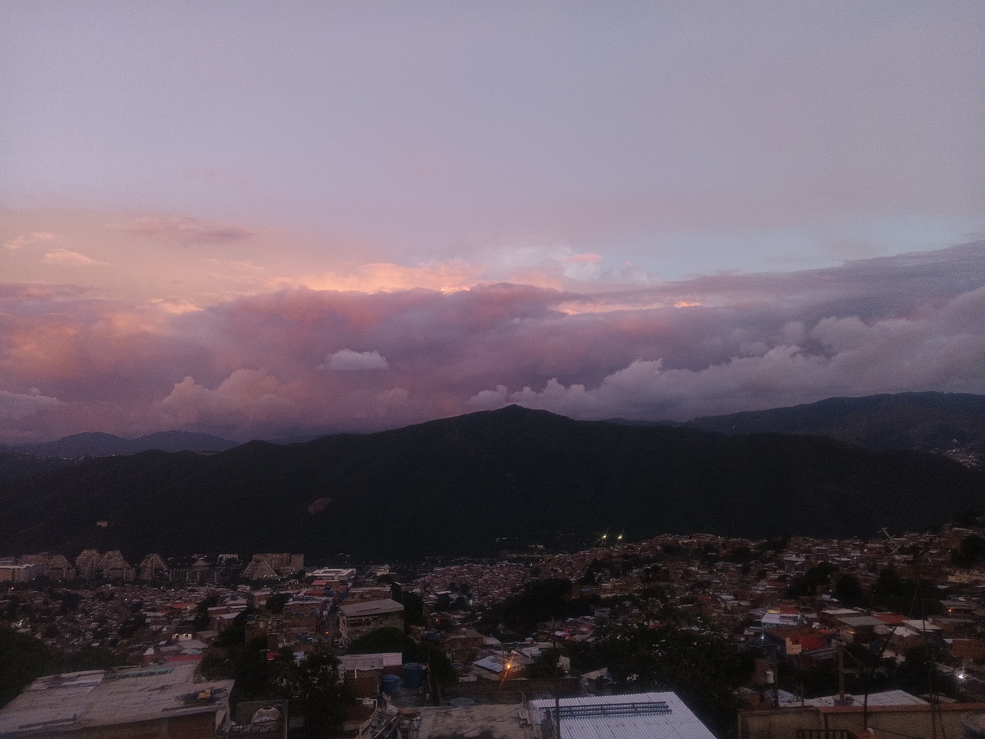 This is an outdoor, medium-shot photograph taken at dusk, looking over a city from an elevated viewpoint. The sky is filled with soft pink and purple clouds. In the background, dark, silhouetted mountains rise against the twilight sky. Below the mountains, a dense urban area stretches across the landscape, characterized by a mix of residential buildings with red-tiled roofs and some taller structures, including what appear to be apartment complexes. Streetlights are visible, dotting the cityscape with small points of light. The overall impression is a sprawling urban environment nestled amongst natural topography during a tranquil evening. This scene is likely from the El Ávila National Park area, overlooking Caracas, Venezuela.