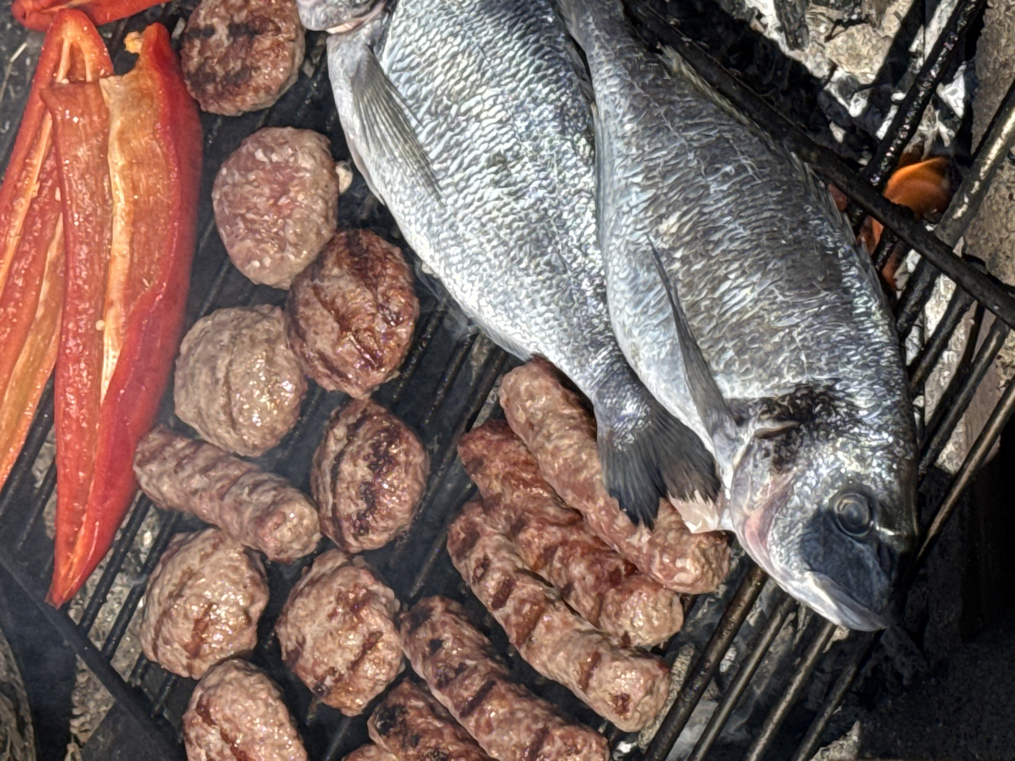 A top-down shot shows a grill with two whole fish, several small sausages, meatballs, and red pepper strips. The fish are silver with dark fins, and they lay diagonally across the grill. The sausages are arranged below the fish, while the meatballs are clustered on the upper left side of the grill. The red pepper strips are also on the left side, next to the meatballs. The grill is made of dark metal bars, and in some spots, flames can be seen beneath the grate. The location appears to be an outdoor cooking area.