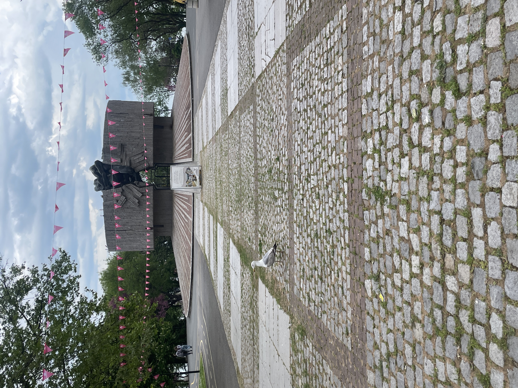 The image shows the Alley of Broken Statues in Burgas, Bulgaria. A seagull stands on a paved walkway leading toward a concrete monument. The walkway alternates between light gray rectangular stones and a darker, smaller cobblestone pattern. Pink triangular flags are strung across the area. Trees and cloudy sky are visible in the background. Two people are seen standing to the lower left, partially obscured by trees.