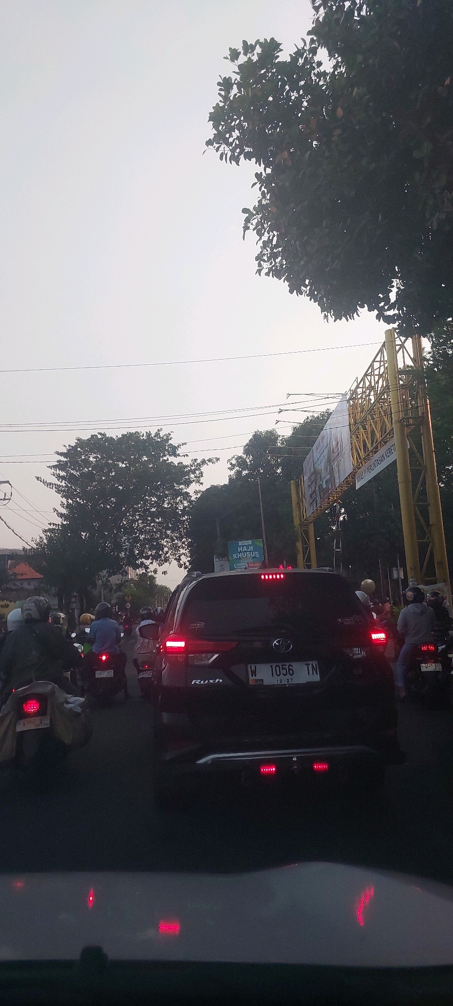 This is a vertical, eye-level shot of moderate traffic on a city street in Surabaya, Indonesia, during daylight hours. The sky is overcast and a light grey. In the foreground, the hood of a car is visible, reflecting red taillight glare from vehicles ahead. Dominating the center of the frame is the rear of a black Toyota Rush SUV. Its taillights are illuminated red, and the license plate reads "W 1056 TN" with a date "12-27". To the left of the SUV, several motorcycles are also visible, some with riders wearing helmets. A yellow metal gantry with advertising banners is present on the right side of the street, with trees and foliage on either side of the road. A sign with "HAJI KHUSUS" is visible in the background. The overall scene depicts a typical traffic jam, likely during a busy commute or rush hour in the urban environment of Surabaya.