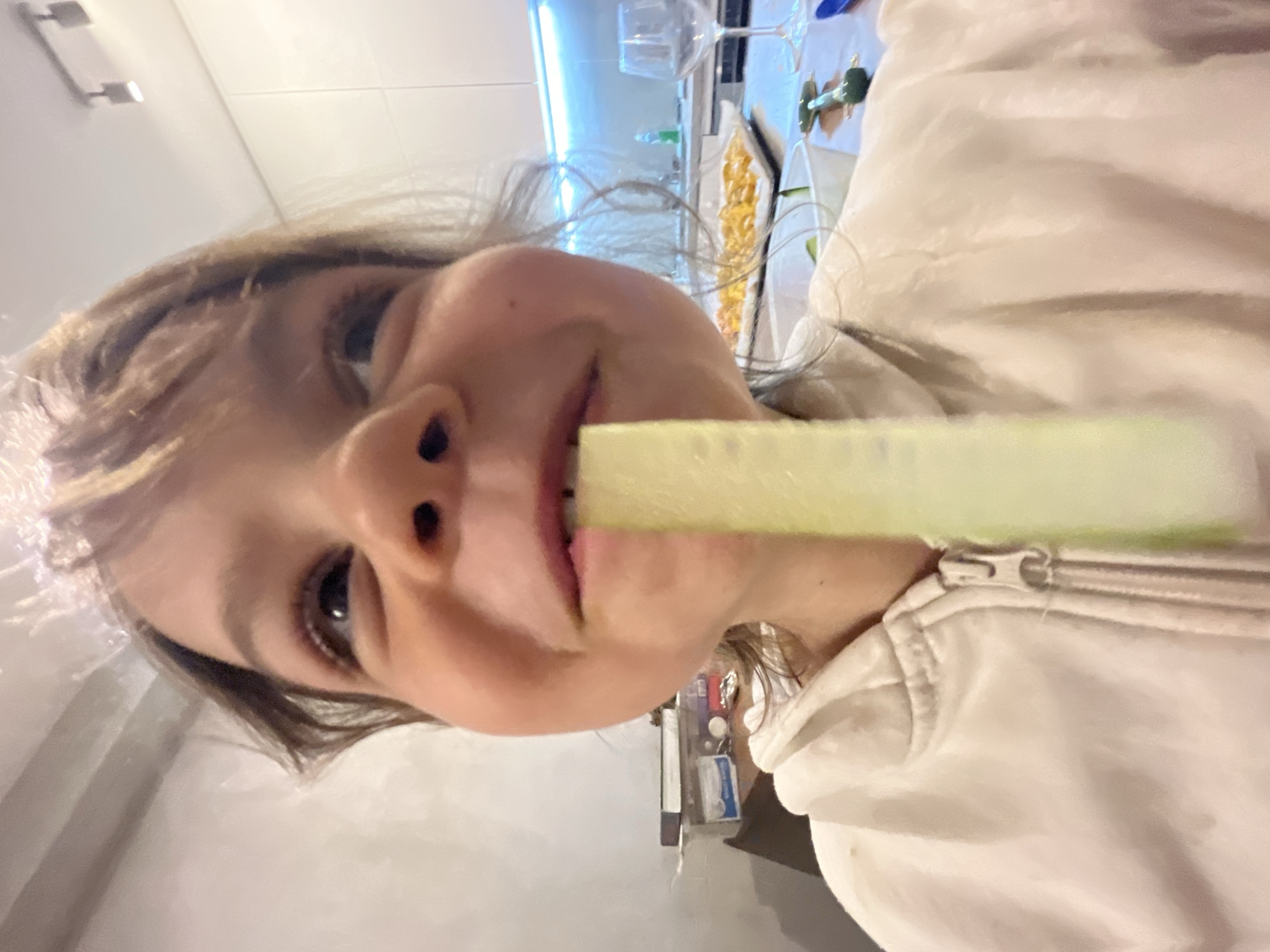 A young child is holding a green cucumber slice in their mouth. The child has light hair and fair skin, and is wearing a white zip-up hooded sweatshirt. In the blurred background are kitchen items, including wine glasses and a tray of food. The location is not discernible from the image and therefore cannot be pinpointed to the city of Budva, Montenegro.