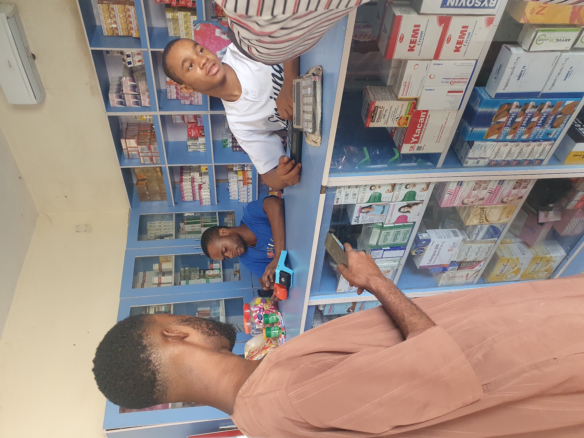 **Location:** Unconfirmed, but presumed to be Gwagwalada, Nigeria.
**People:** The image shows three people. One in a white T-shirt looking at the camera, one wearing a blue T-shirt is looking down towards a display of objects, and one in a peach/brown shirt is holding a phone.
**Subjects:** The main subject is a retail or pharmacy environment.
**Objects:** There are shelves stocked with various pharmaceutical products and medications. There is also a small display of colorful children’s toys or candies. Other objects include the mobile phone and electronic payment device.
**Actions:** The person in peach/brown shirt is holding a phone, possibly taking a picture, while the person in the blue t-shirt seems to be interacting with the toys. The person in the white T-shirt is looking directly at the camera, possibly assisting customers.
**Interactions:** Presumed interaction is between a customer, a store worker, and their phone.
**Environment:** The environment is a pharmacy or retail space. The walls are lined with shelves filled with merchandise. Lighting appears to be a combination of natural and artificial light.
