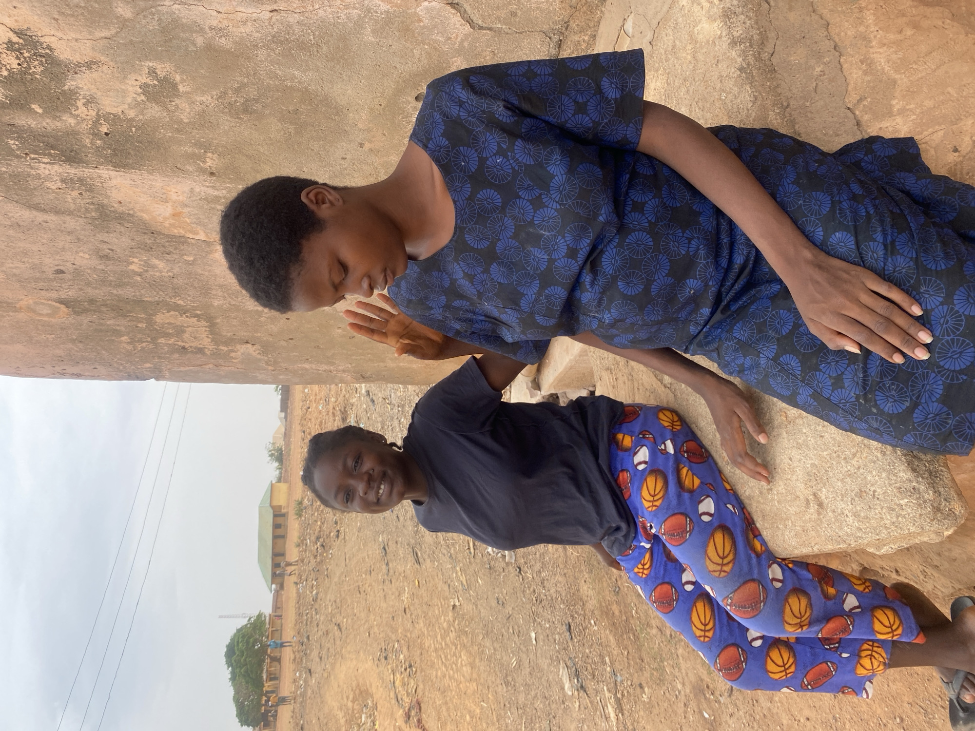 Two young women are sitting on a stone in an outdoor setting. One woman wears a blue and black patterned dress and has short, dark hair. She appears to be looking down. The other woman wears a dark T-shirt and blue pants patterned with images of sports balls. She is smiling. The setting appears to be in a rural area, possibly on the outskirts of Rido, Nigeria, based on the landscape and building style visible in the background.