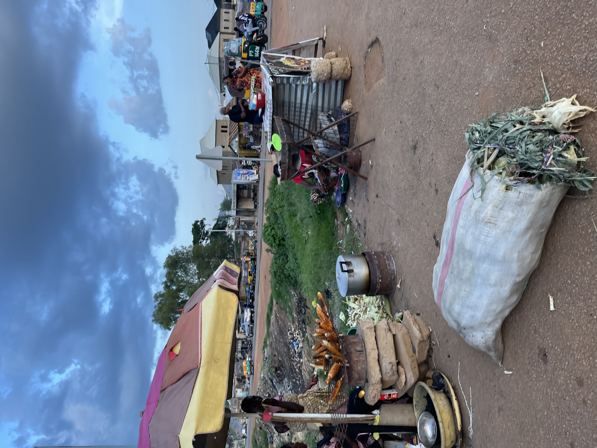 This image depicts a bustling street scene in Jos, Nigeria, centered around roadside vendors and daily activities under an overcast sky.
In the foreground, a vendor, likely a woman, is seen seated on the ground, attentively tending to a metal grill laden with several ears of roasting corn. She is dressed in vibrant, patterned attire. Adjacent to her setup is a large metal pot with a lid, resting on a cylindrical base, and a stack of light-colored bricks, possibly used for cooking. Two sacks are also present: one large grey sack with a pink stripe, filled with green plant material (likely corn husks), and another burlap sack containing more corn. To the left, a large, multi-colored canopy with stripes of pink, yellow, and brown offers shade.
Further back, a makeshift stall fashioned from corrugated metal sheets is visible, behind which another individual, possibly an older man with grey hair, stands, appearing to interact with customers. Along the dusty road in the mid-ground, several three-wheeled auto-rickshaws (keke napeps) and other vehicles are parked. One keke napep clearly shows the license plate "PF 166ZE". Beyond the road, a line of light-colored buildings with dark roofs can be seen, indicative of a semi-urban environment. Between the stalls and the road, a stretch of unkempt ground or a trash heap is noticeable.
The sky is heavily cloudy, featuring a mix of dark grey and lighter blue tones, suggesting an overcast day or the onset of evening. Patches of green grass and trees are scattered throughout the scene. The image portrays a slice of everyday life and commerce in a Nigerian street market.