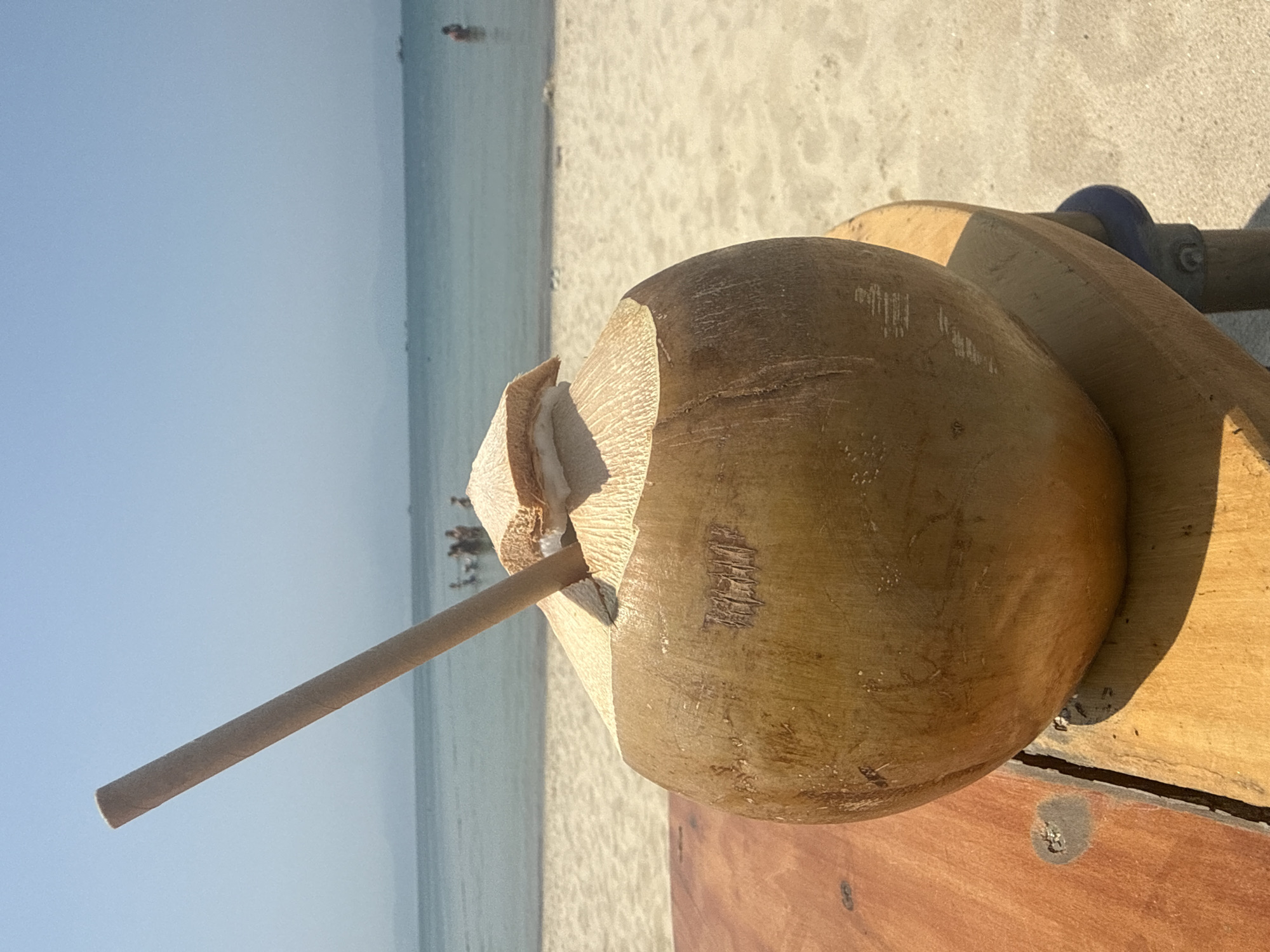 A light brown, partially husked coconut, with its top section removed to expose white flesh, occupies the foreground on the left. A single light brown cylindrical straw is inserted into the coconut's opening, angled upward to the left. The coconut rests on a multi-planked, worn wooden surface that exhibits grain and scratches. A shadow of the coconut is cast onto this surface, indicating strong sunlight from the right.
Behind the coconut and wooden surface, a wide expanse of light-colored sand extends across the midground, forming a beach. Beyond the sand, the calm, light blue-green ocean fills the background. Several individuals are visible in the water; a group of approximately five people are clustered on the left side, and a single person is located further right. In the far distance, the horizon shows faint outlines of landmasses, consistent with mountains or hills, under a clear, light blue sky. Small, dark, indistinct objects, possibly vessels or buoys, are visible on the water toward the horizon. The scene depicts a sunny day at a coastal beach in Đà Nẵng, Vietnam.